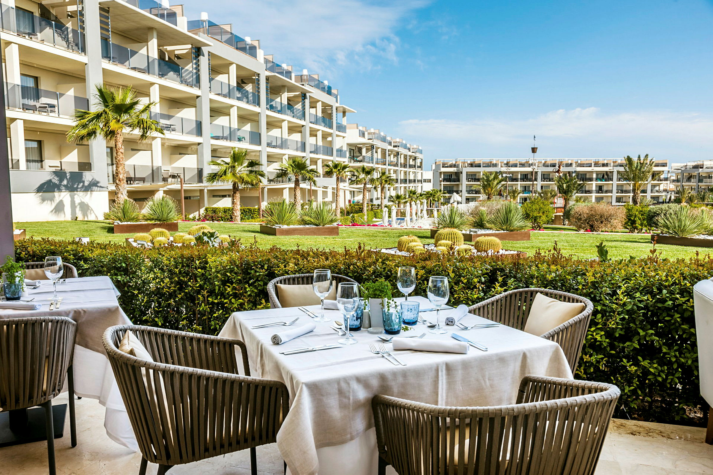 Bilde av en utendørs restaurant med dekkede bord, omgitt av en hage og moderne bolig i bakgrunnen. (AI-generert tekst)