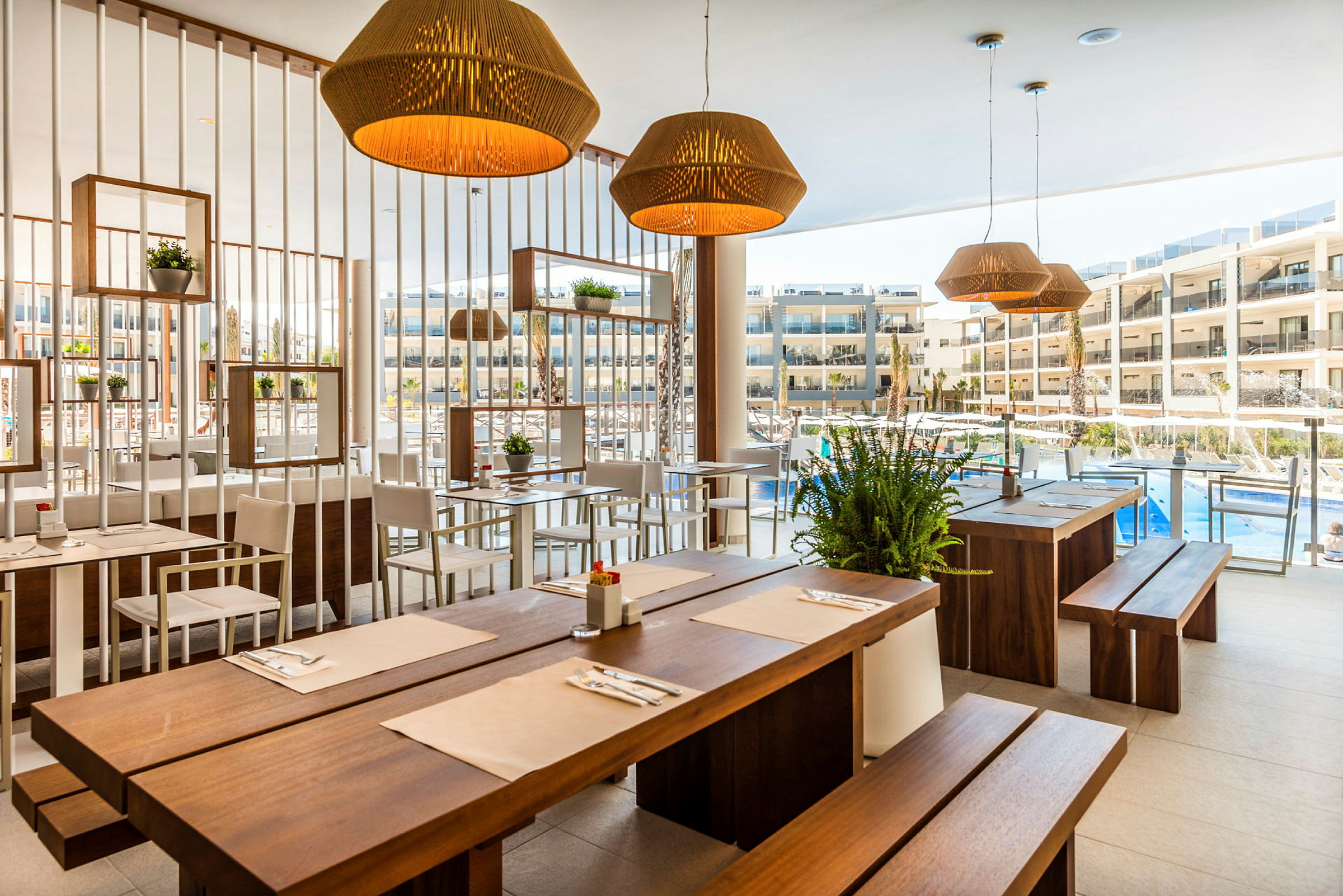 Restaurant med trebord og lyse stoler, dekorert med planter og lamper, med utsikt mot et basseng og hotellbygninger. (AI-generert tekst)