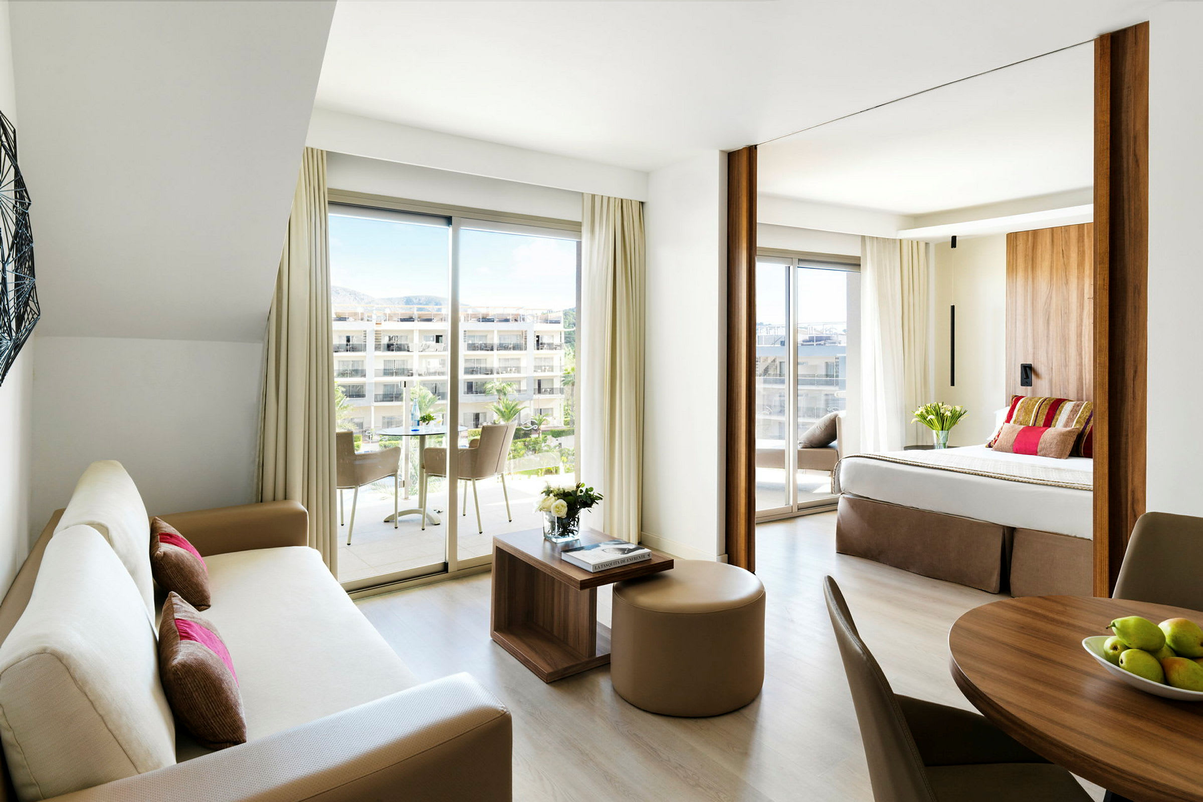 Et moderne hotellrom med åpen planløsning, inkluderer en stue med sofa og et soverom med dobbeltseng samt store vinduer med utsikt mot en terrasse. (AI-generert tekst)