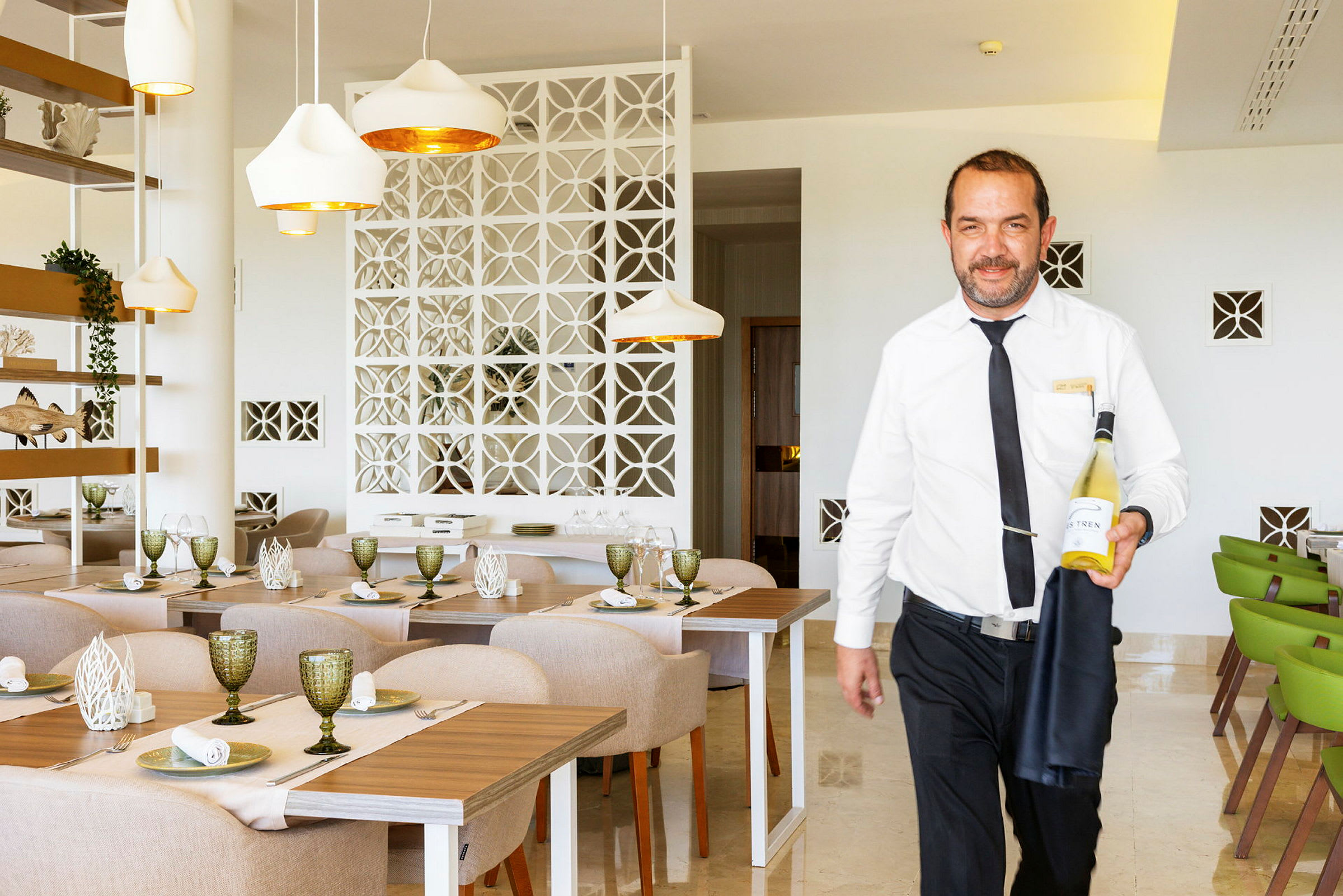 En kelner i hvit skjorte holder en flaske vin i en lys og moderne restaurant. (AI-generert tekst)