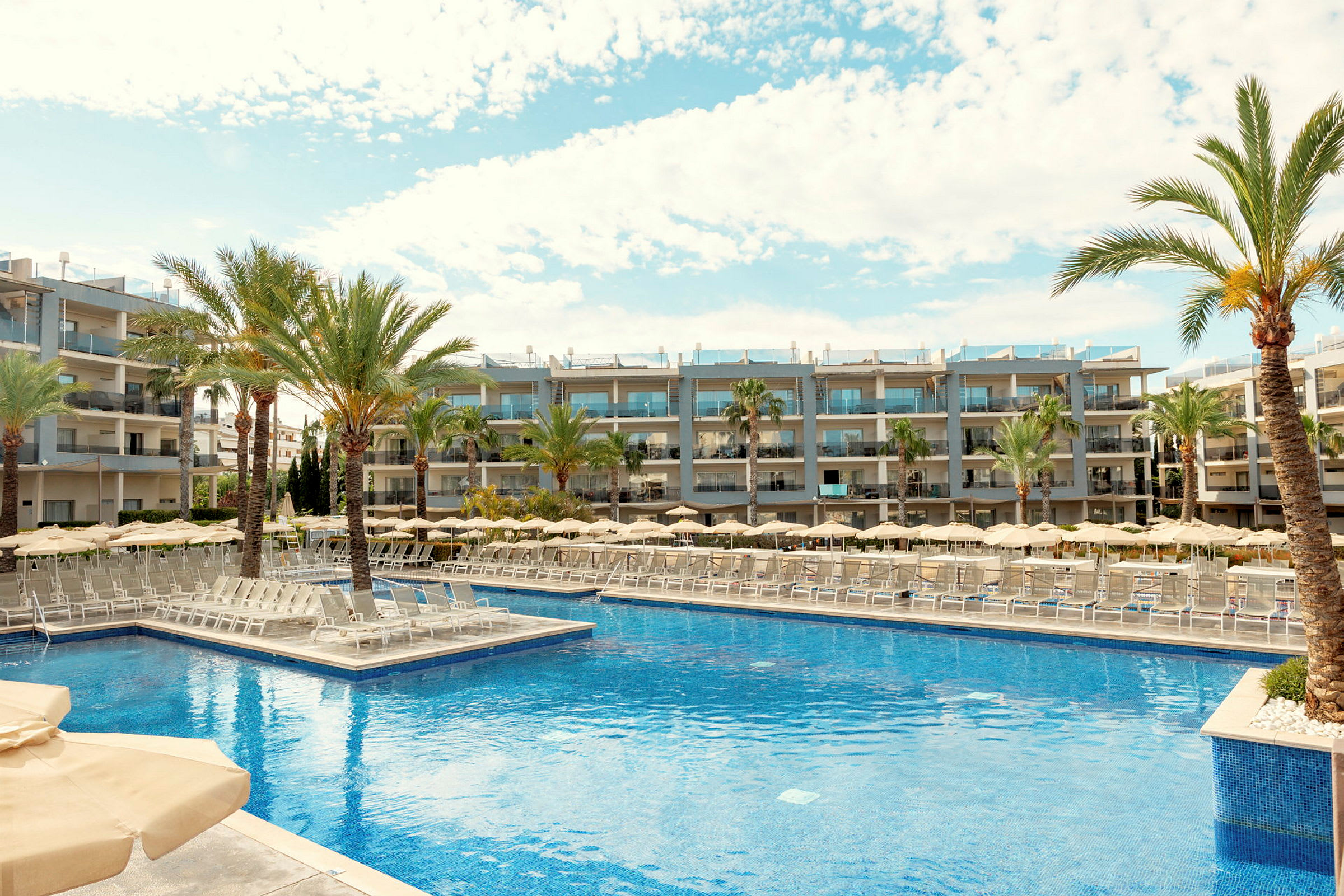 Et bilde av et hotellområde med et stort basseng omgitt av solsenger, parasoller og palmer. I bakgrunnen ses moderne hotellbygninger. (AI-generert tekst)