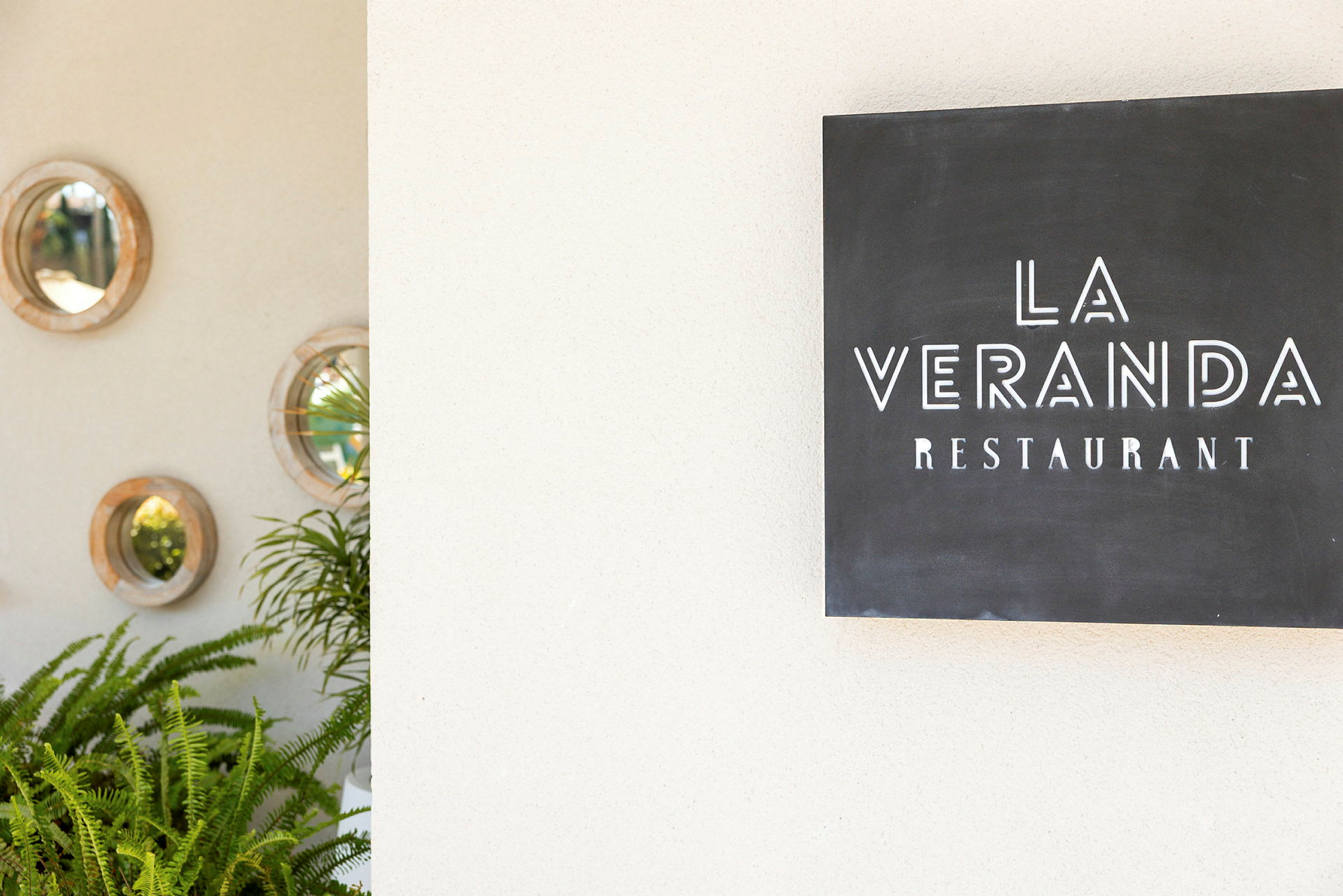 Et skilt som viser teksten "La Veranda Restaurant" ved siden av en vegg dekorert med runde speil og grønne planter. (AI-generert tekst)
