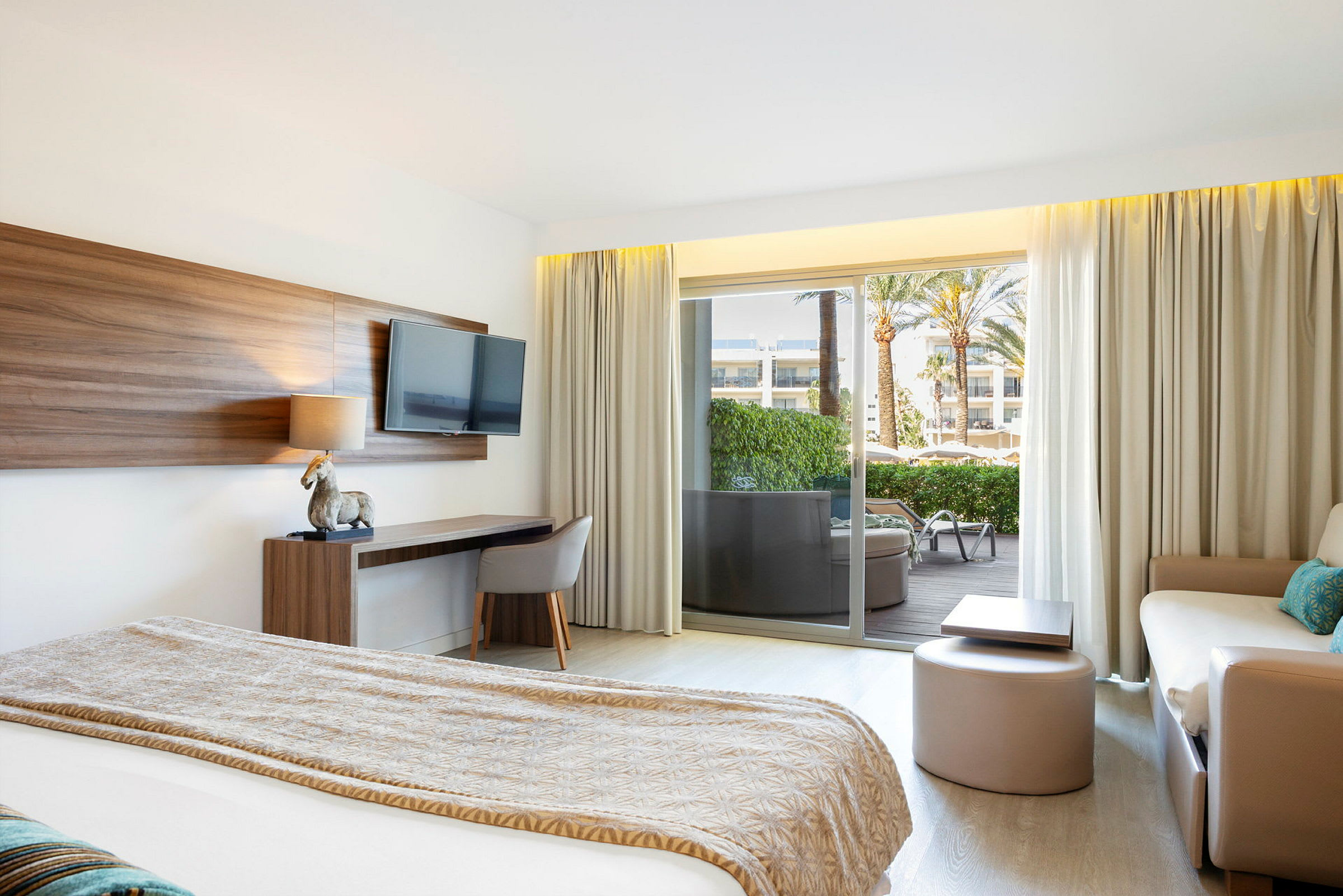 Moderne hotellrom med en seng, sofa, arbeidsbord og en TV på veggen. Glassdør mot en balkong med utsikt over en hage. (AI-generert tekst)