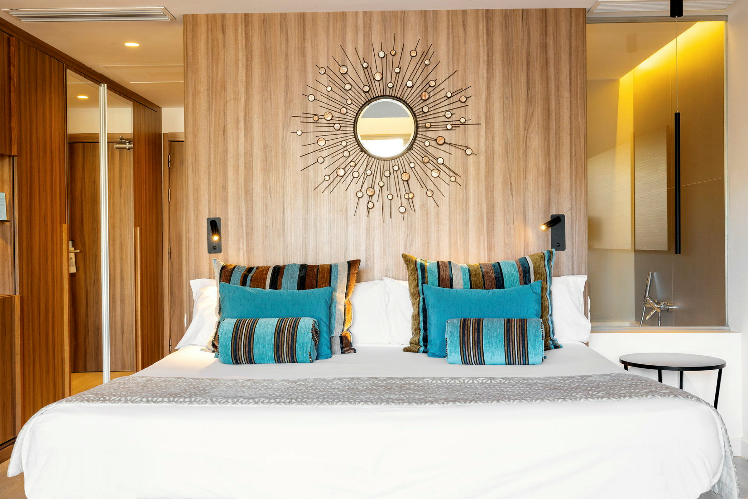 Et hotellrom med en stor seng dekorert med fargerike puter i blått og stripete mønster, en trevegg med et speilmotiv og moderne belysning. (AI-generert tekst)