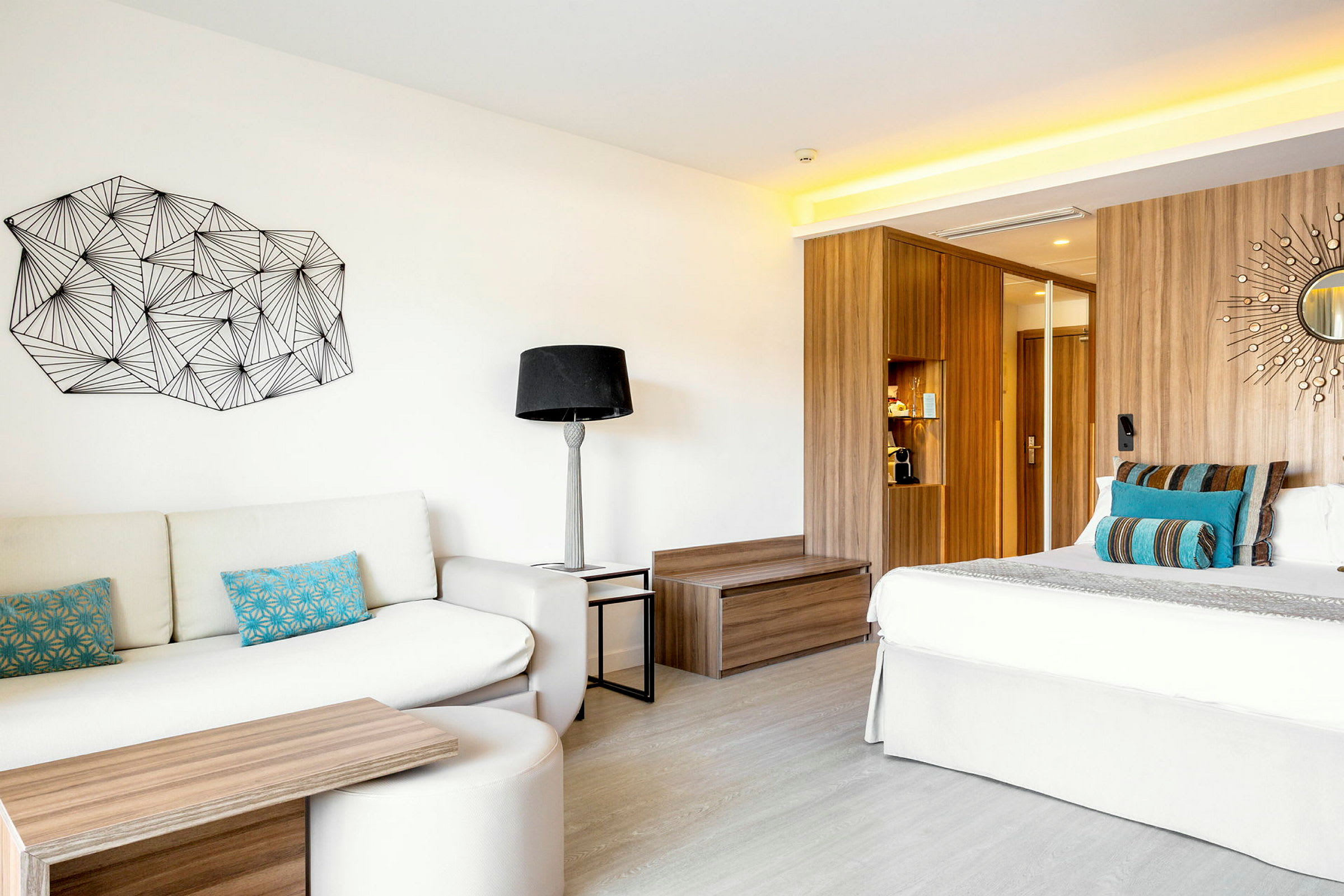 Et moderne hotellrom med en sofa, fargerike puter, et sofabord, en seng med dekorative puter og en trepanel bak. På veggen er det kunst og en liten rundt speildekorasjon. (AI-generert tekst)