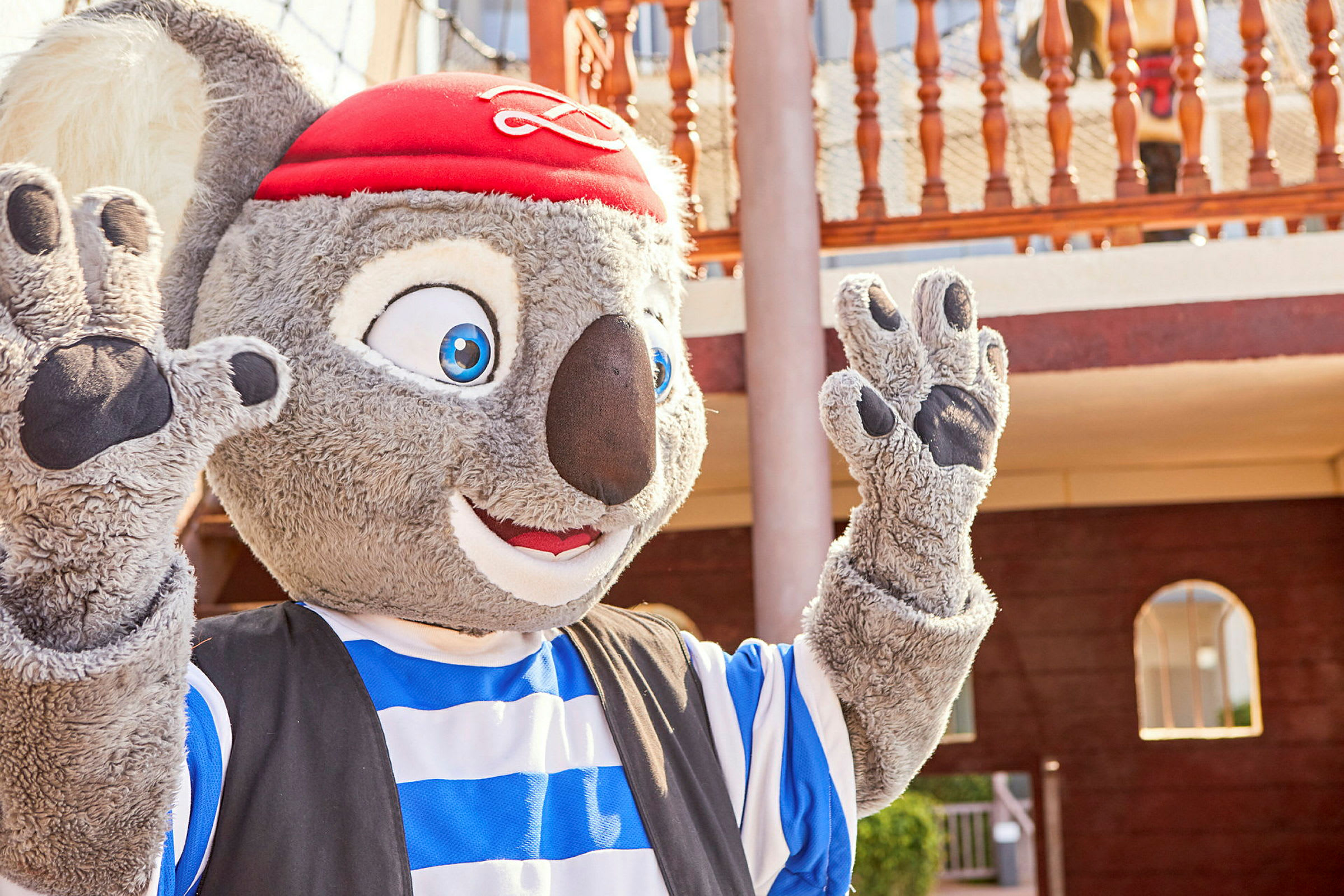 Maskot med grå pels, stripet blå og hvit drakt samt rød hatt, synlig foran en trebalkong. (AI-generert tekst)