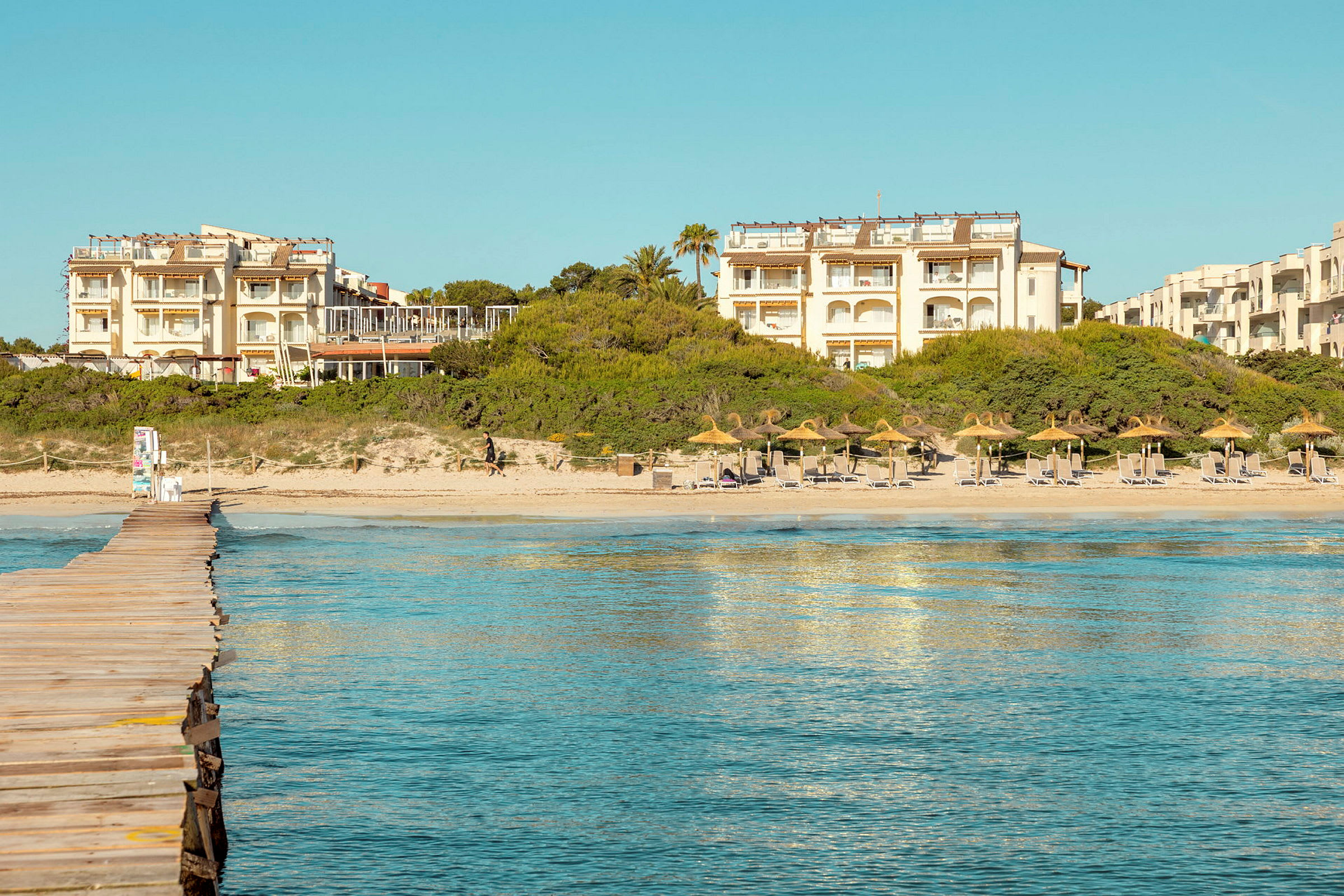 En strandpromenade leder til en sandstrand med parasoller og solsenger. I bakgrunnen er det hotellbygninger og vegetasjon. (AI-generert tekst)