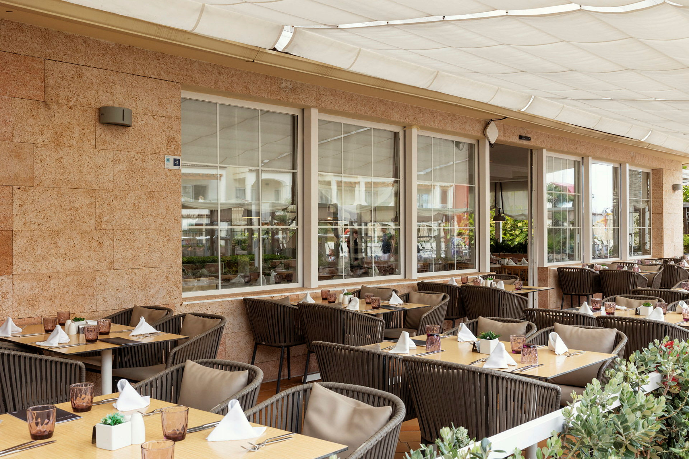 En utendørsterrasse ved en restaurant med dekkede bord og stoler, omgitt av grønne planter. (AI-generert tekst)