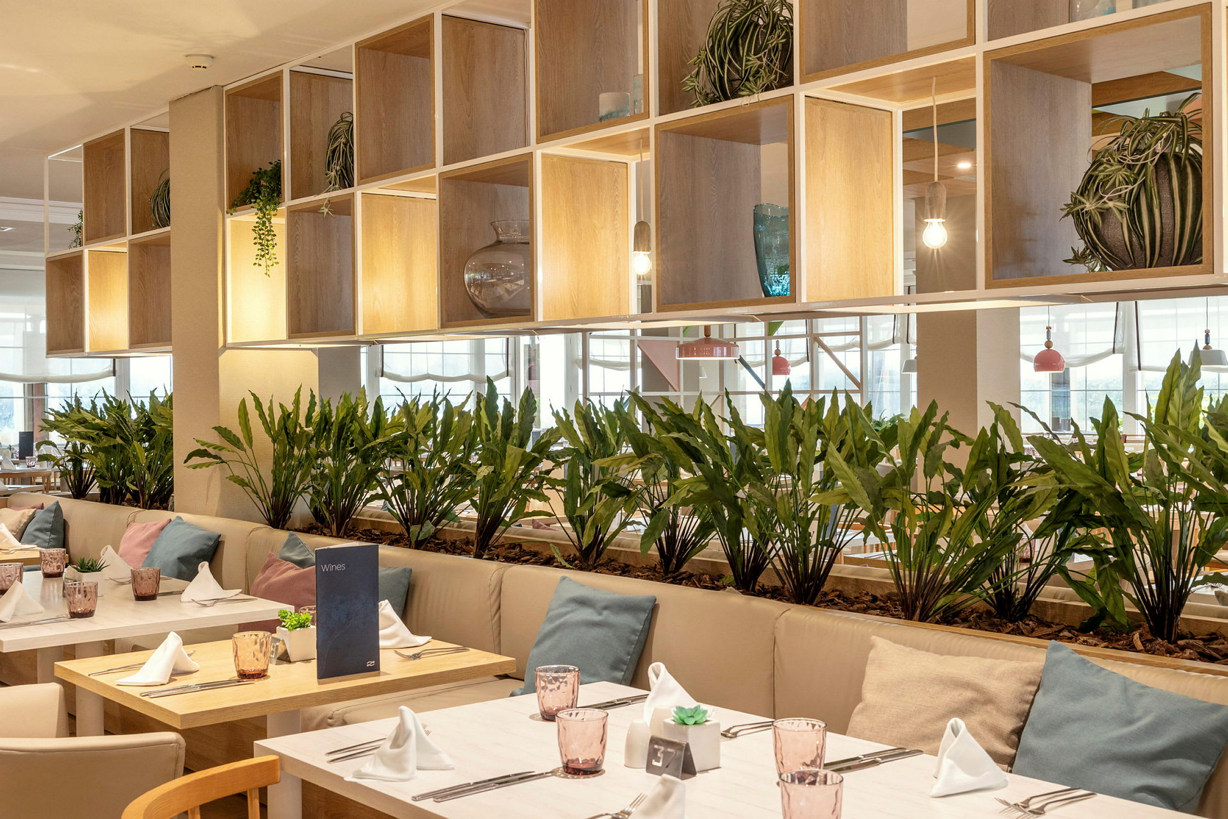 Et moderne dekorert restaurantområde med bord pyntet med hvite servietter, glass og puter på sofaene. Grønne planter plassert som skillevegger. (AI-generert tekst)