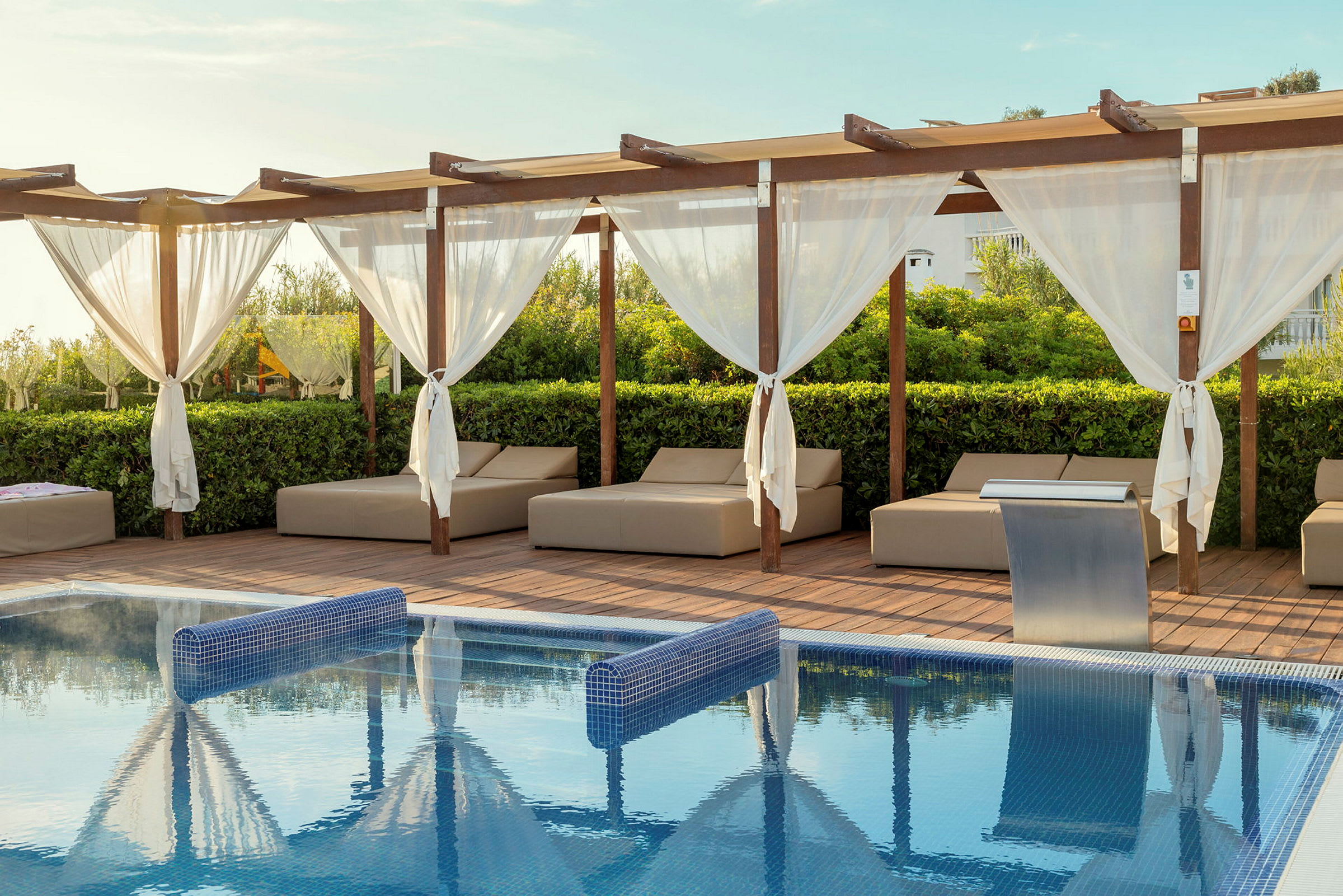 Et basseng med blått vann, omgitt av solsenger og en terrasse med pergola og hvite gardiner. (AI-generert tekst)