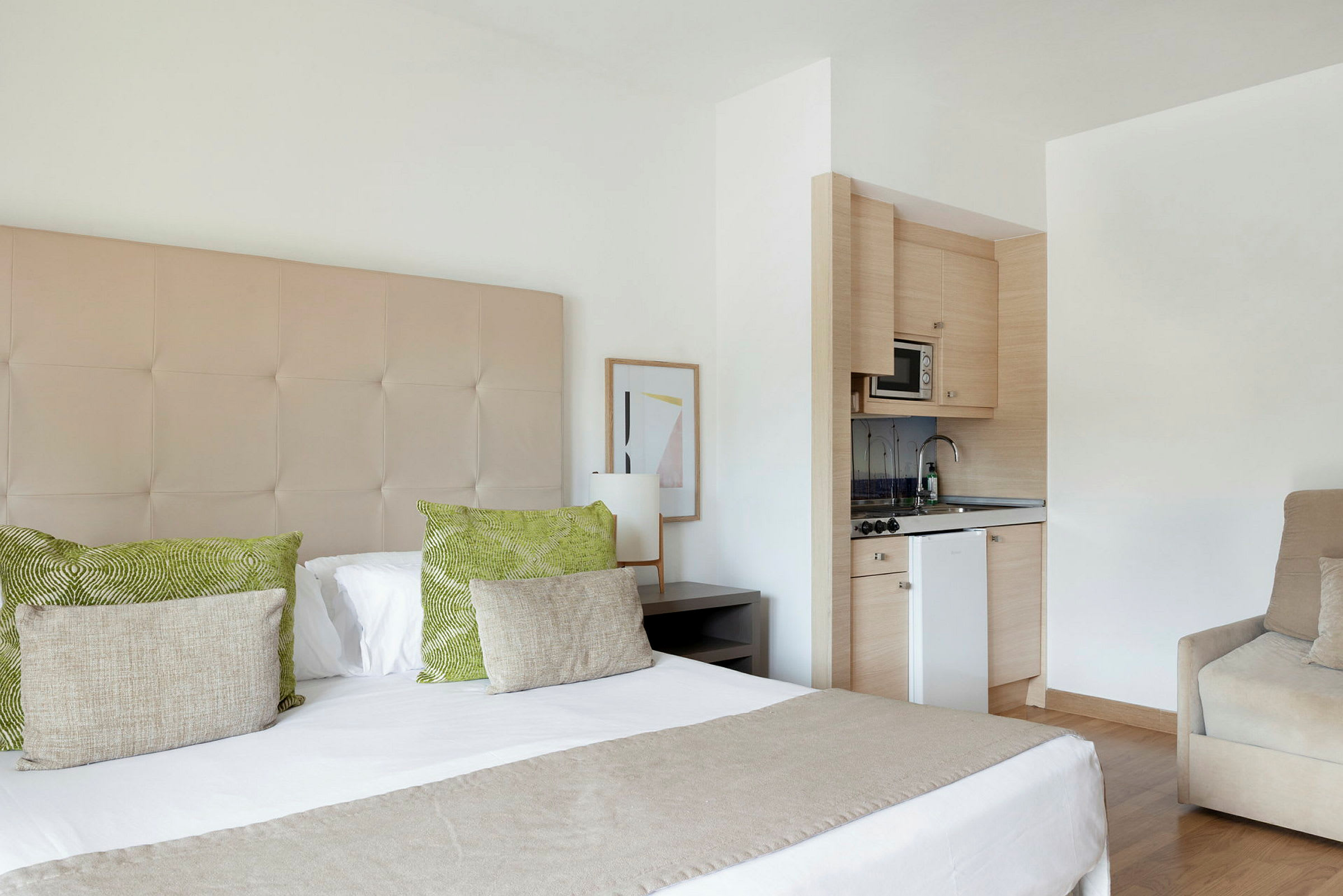 Et hotellrom med en dobbeltseng, grønne og beige puter, en liten kjøkkenkrok med mikrobølgeovn og vask samt en beige lenestol. (AI-generert tekst)