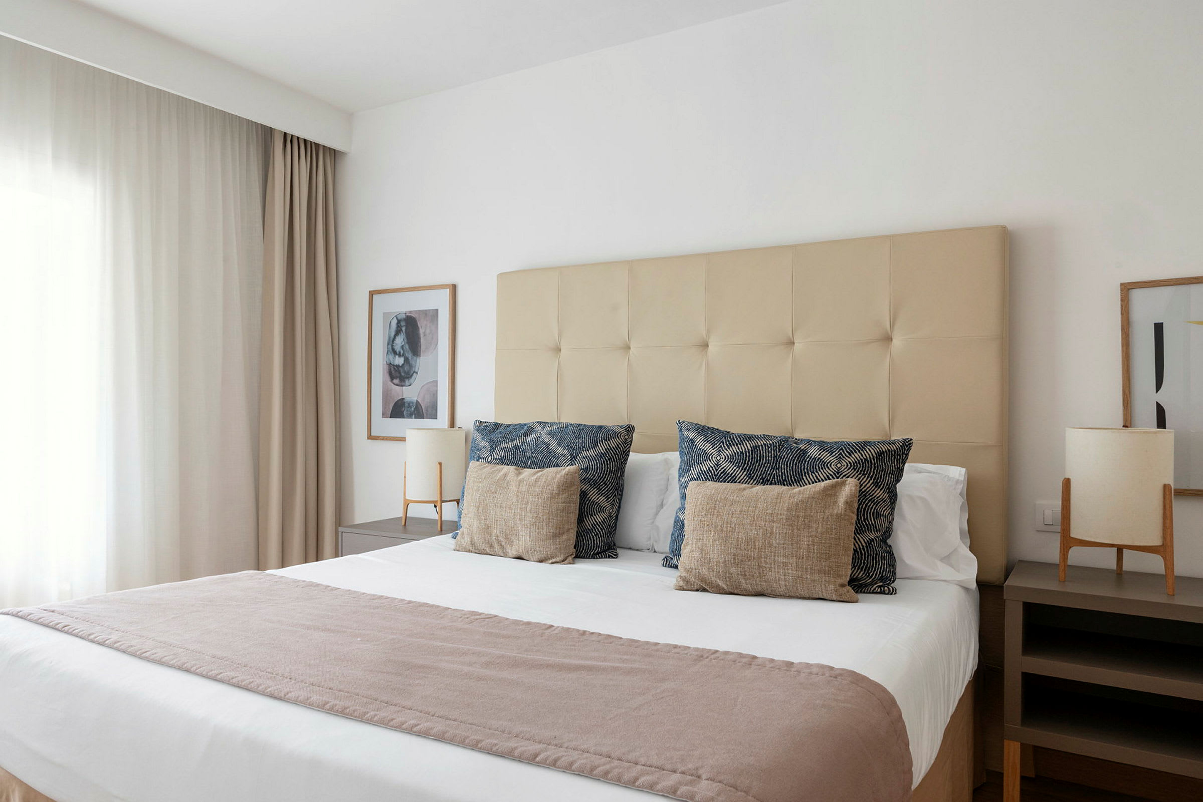 Et hotellrom med en stor seng dekket av hvite laken, dekorative puter og et lyst teppe. Sengen er plassert mellom to nattbord med lamper. (AI-generert tekst)