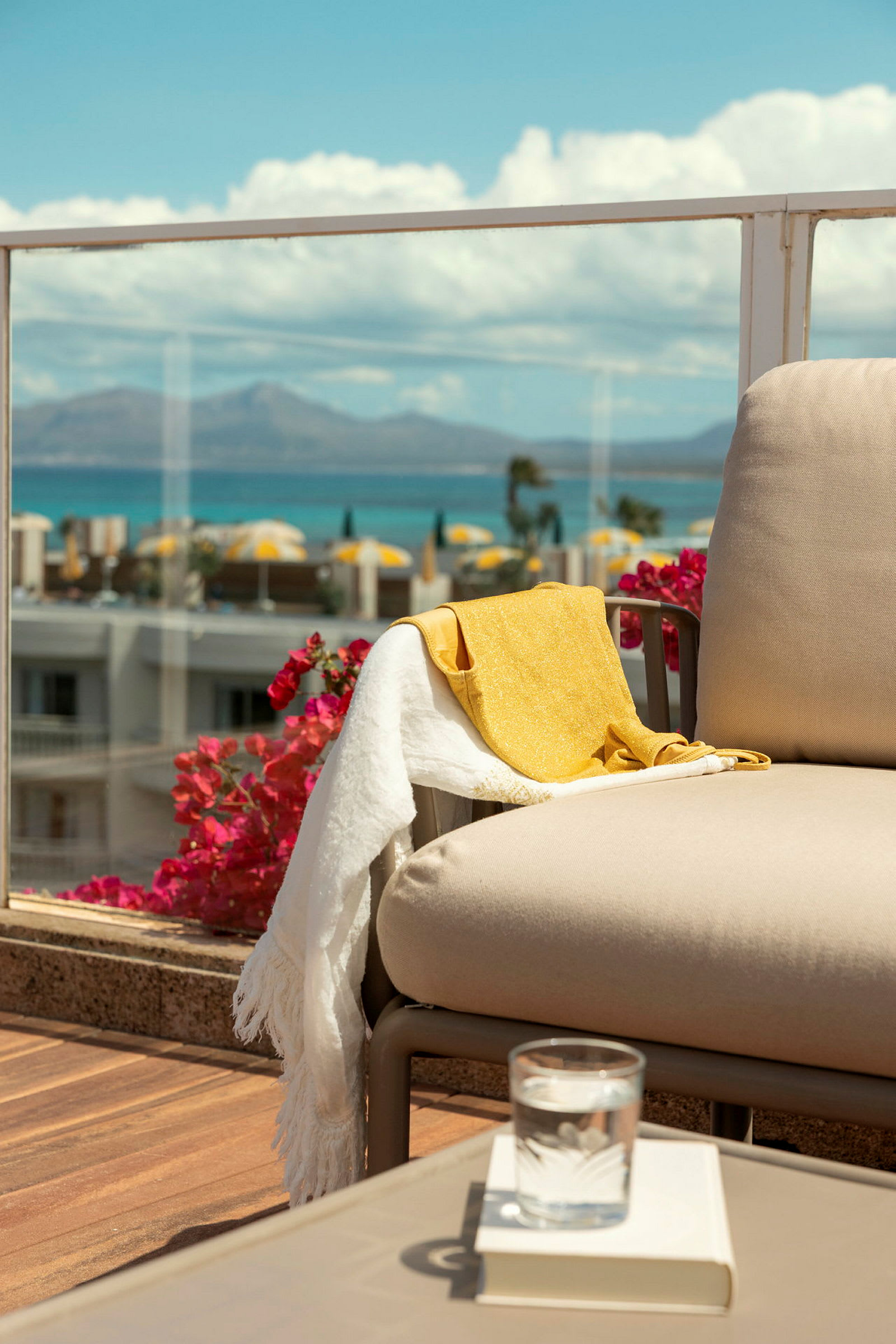 En beige stol på en terrasse med utsikt over havet og det omkringliggende landskapet, med et gult og hvitt håndkle hengende over armlenet. (AI-generert tekst)