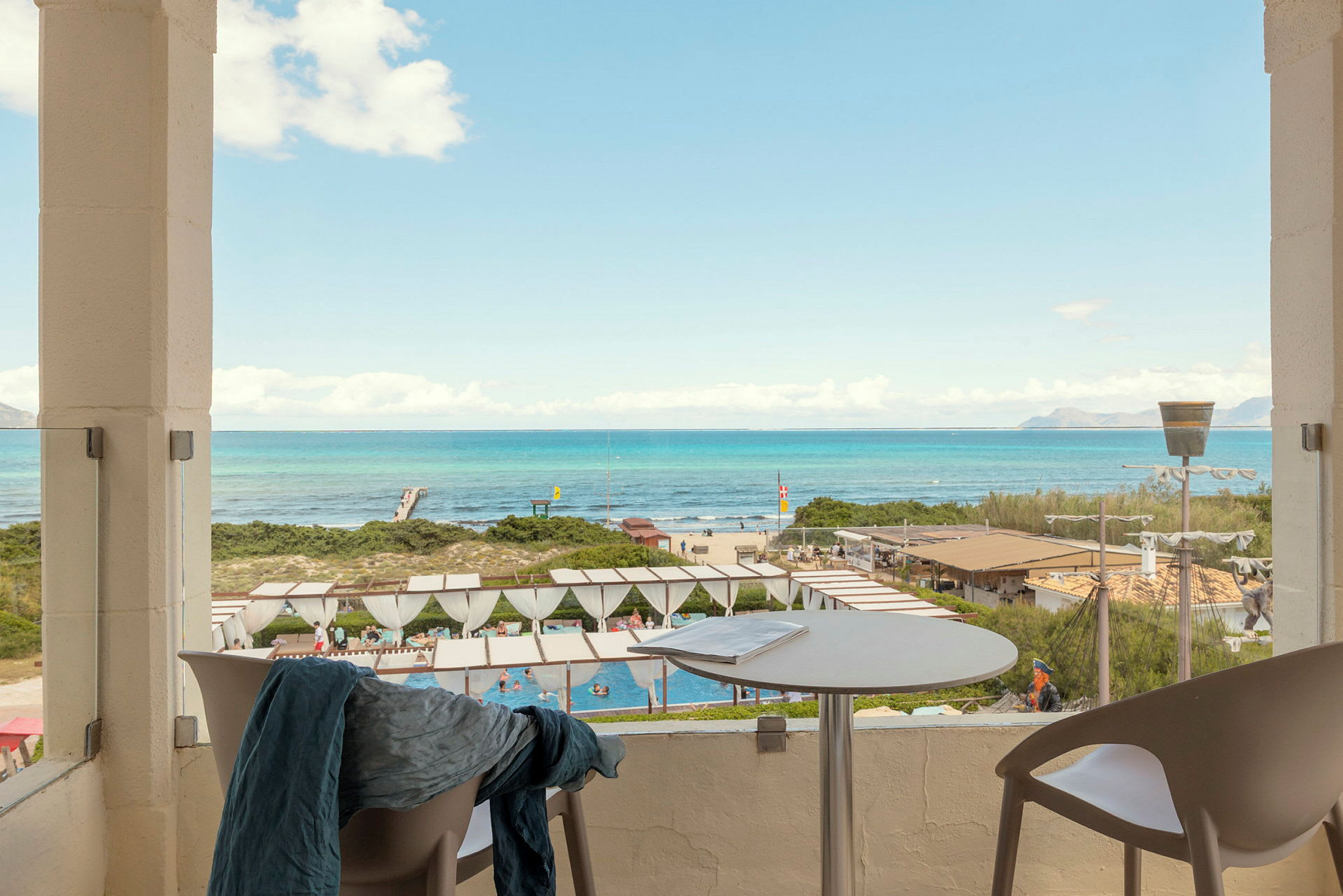 Utsikt fra en uteplass mot et bassengområde og en strand med turkist hav i bakgrunnen. (AI-generert tekst)