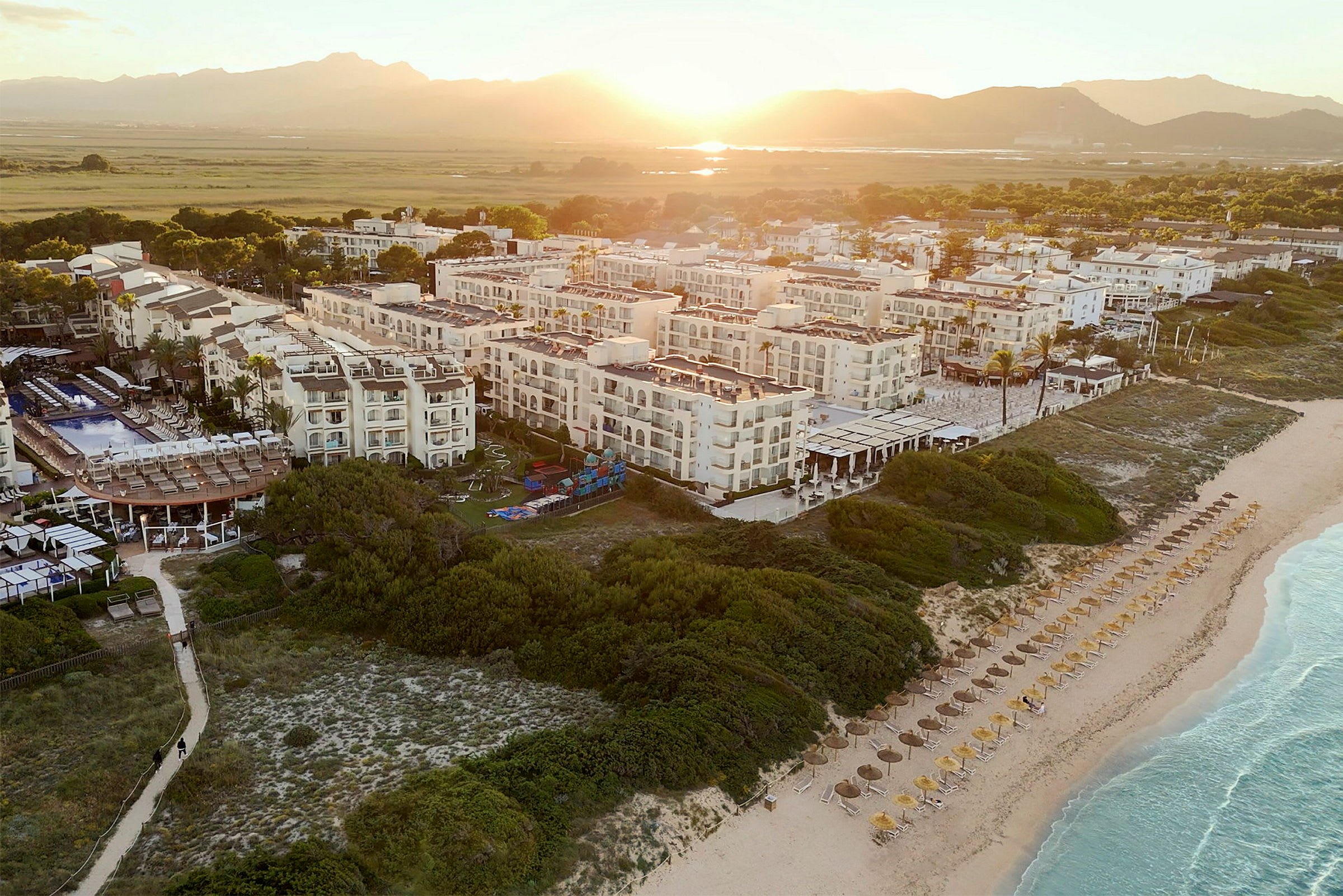Luftfoto av et hotellkompleks ved stranden, omgitt av grøntområder og med soloppgang i bakgrunnen. (AI-generert tekst)