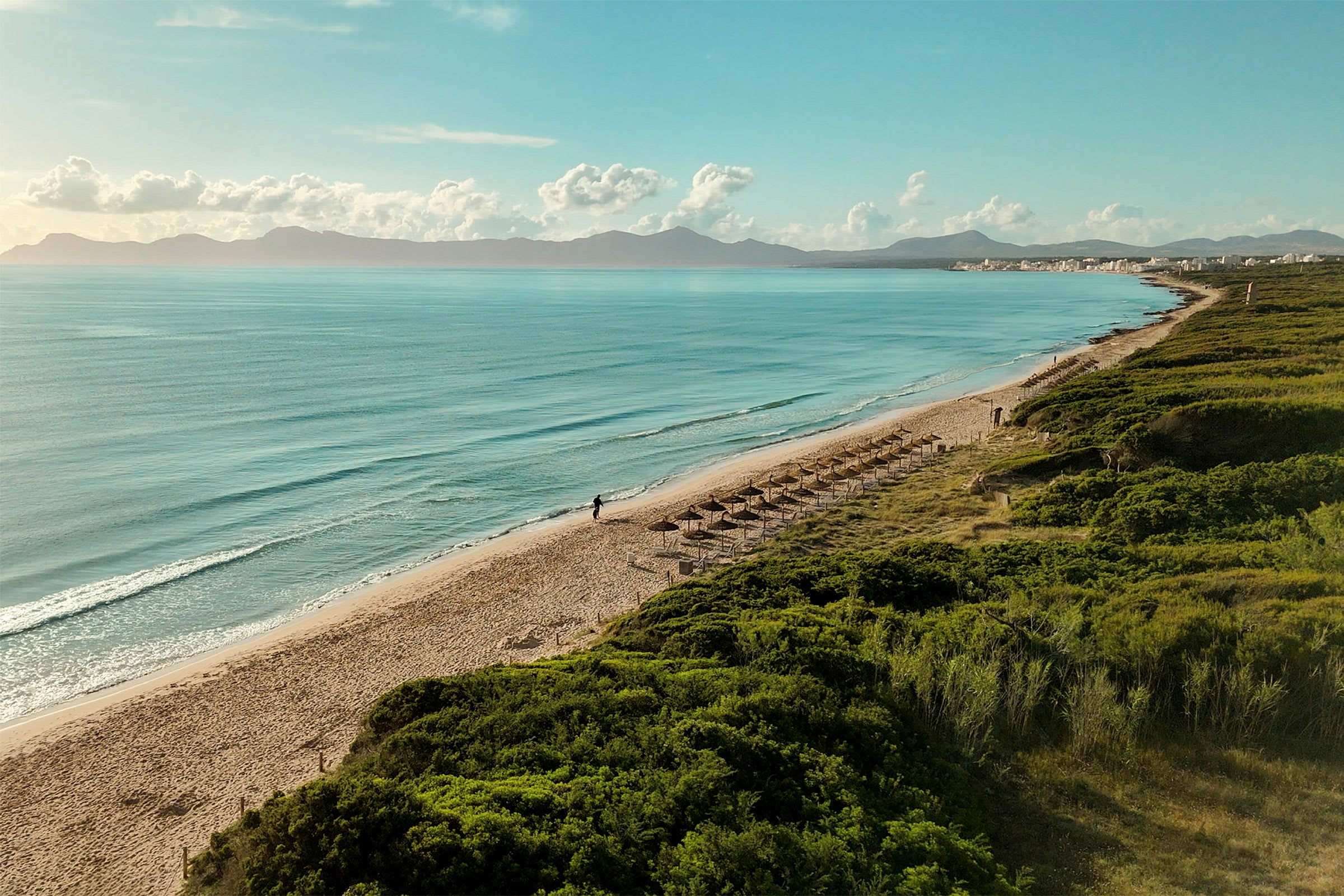 Strand med parasoller, sjø og fjell i bakgrunnen. (AI-generert tekst)