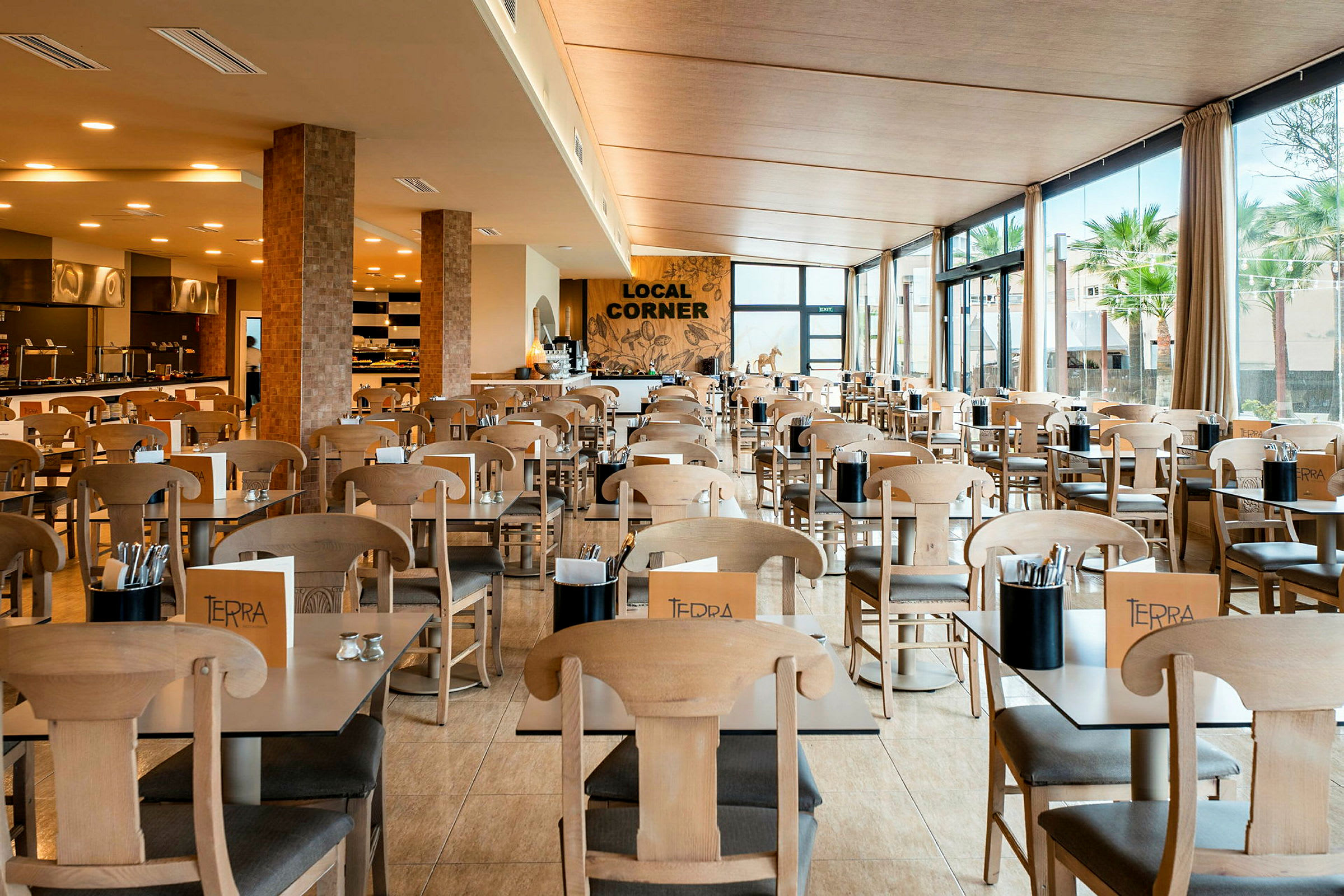 Et lyst og romslig restaurantområde med bord og stoler i tre. Store vinduer med utsikt over grønne omgivelser. (AI-generert tekst)