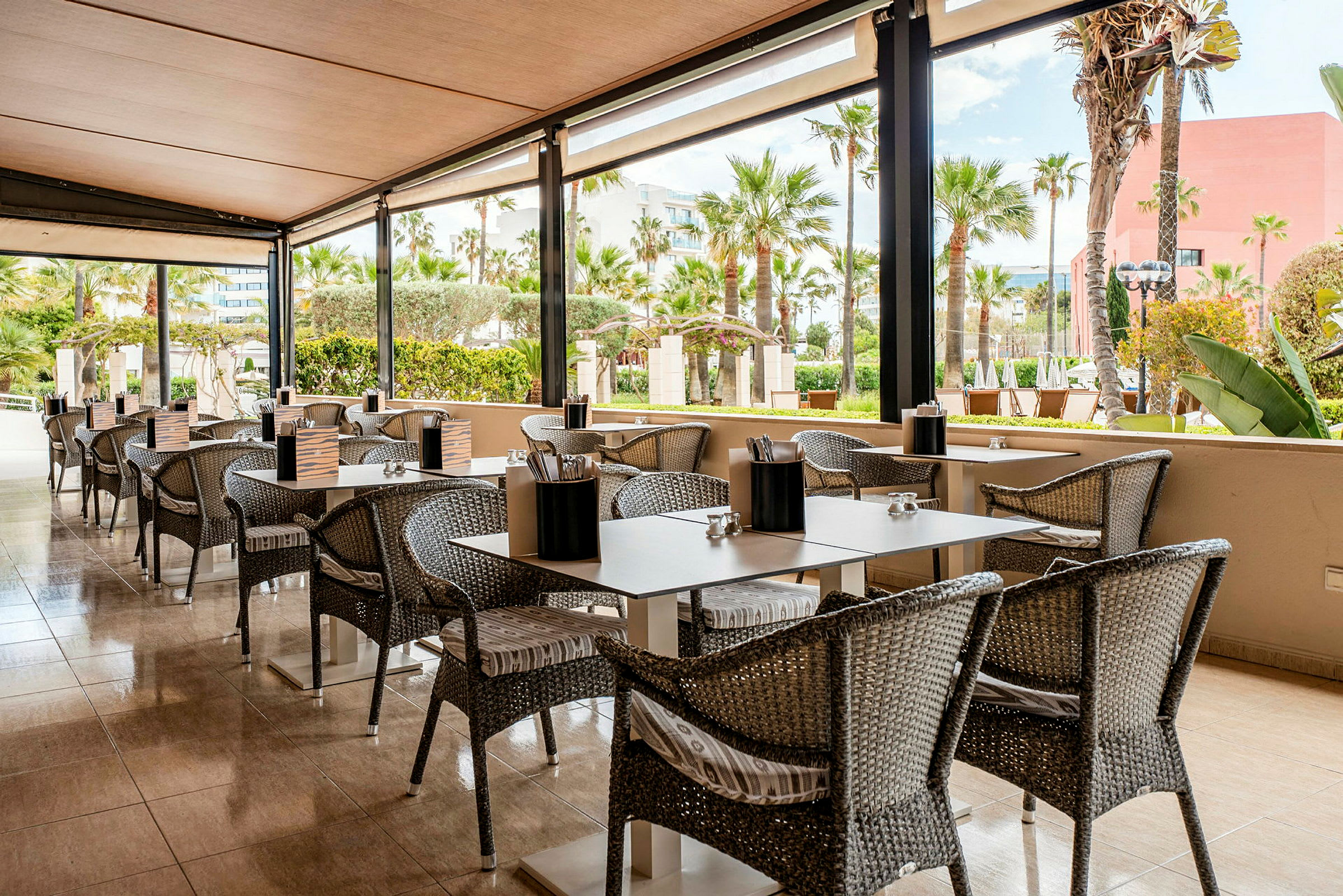 En utendørs restaurant med bord og stoler på en terrasse med utsikt over en hage med palmer. (AI-generert tekst)