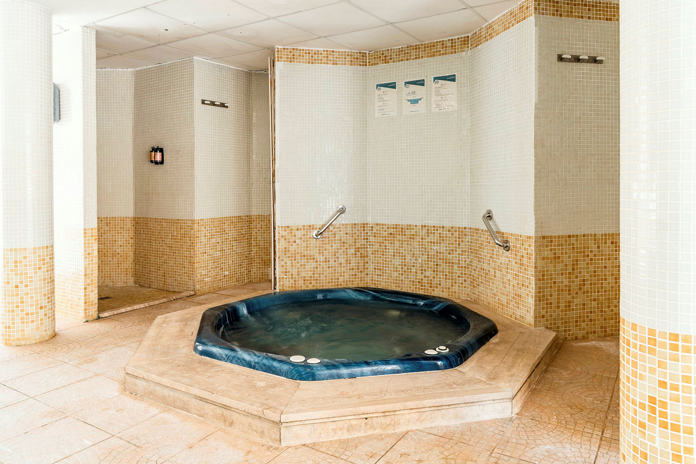 Bildet viser et rom med et boblebad omgitt av mosaikkdekor på veggene. Boblebadet er blått og nedsenket i gulvet. (AI-generert tekst)