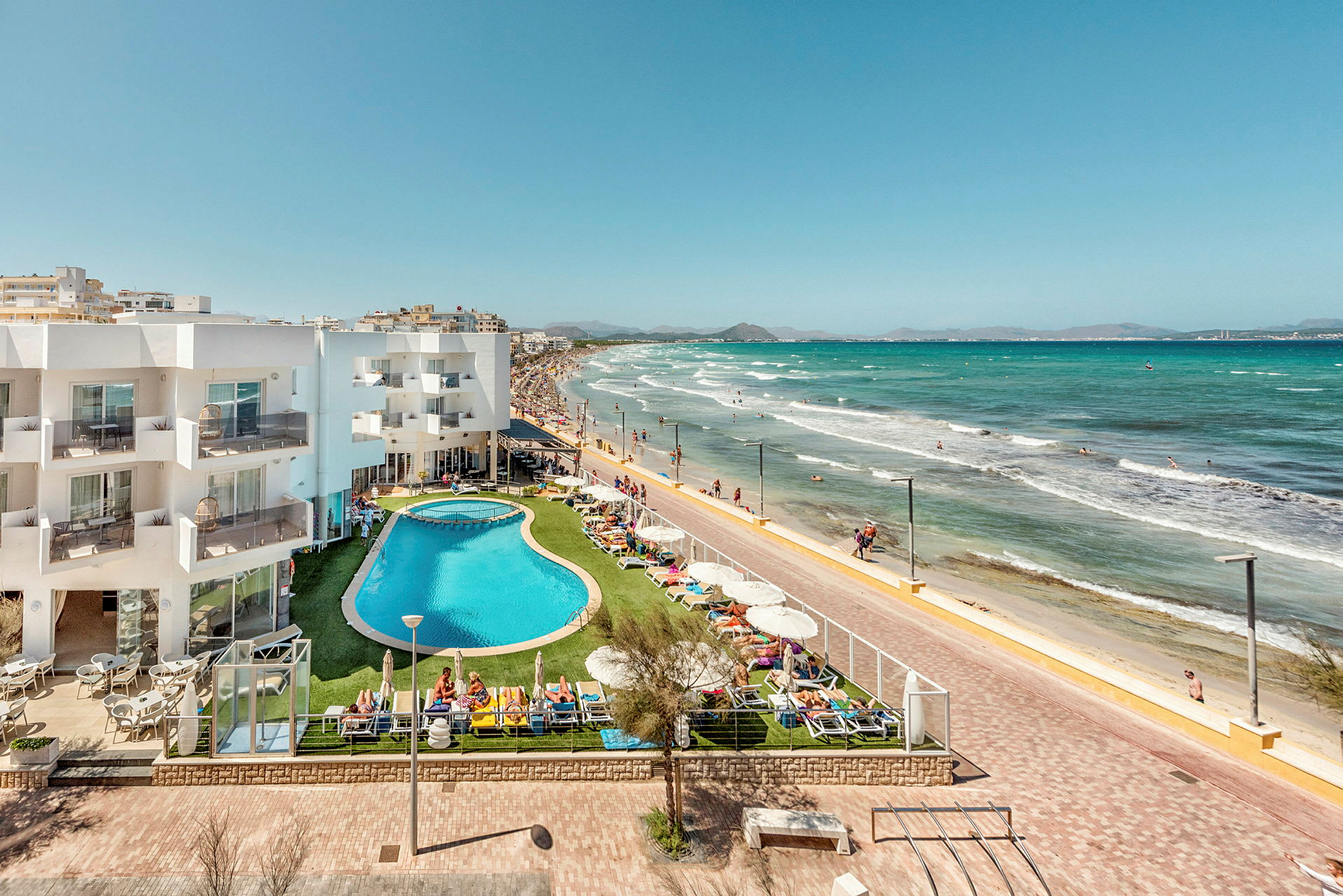 Hotellbygning og bassengområde ved strandpromenade og sandstrand. (AI-generert tekst)