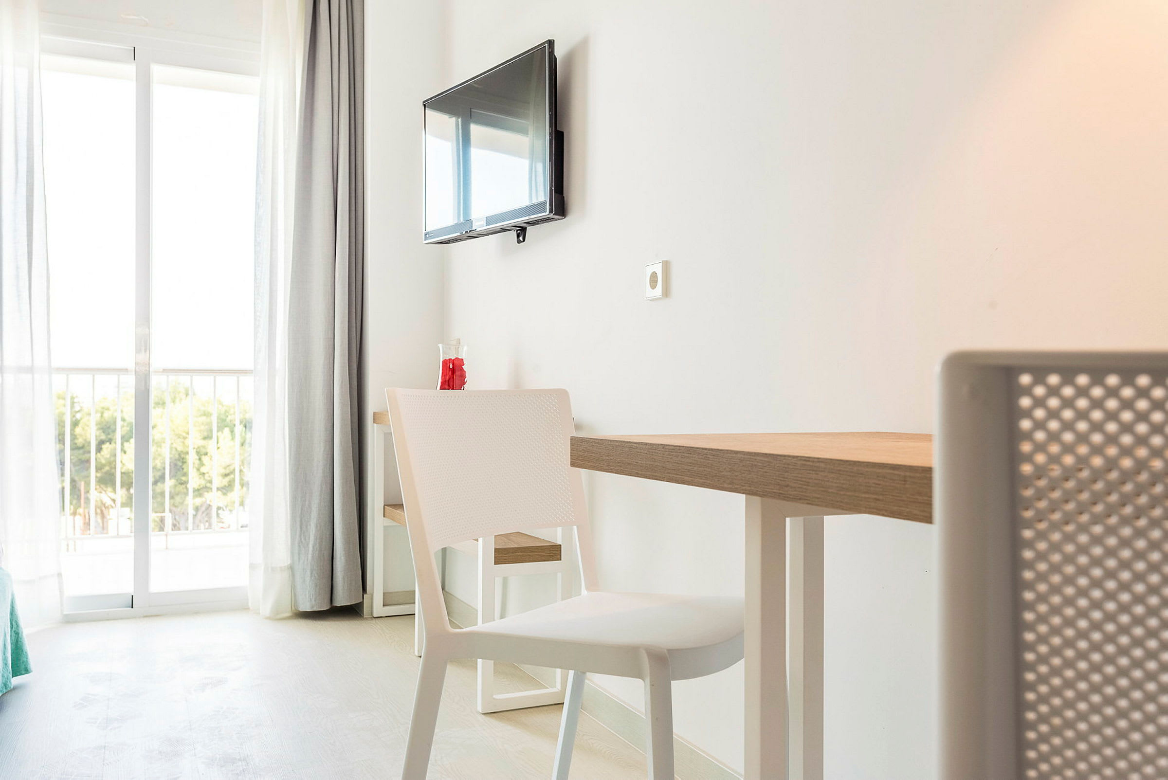 Et hotellrom med hvitt interiør, et bord og stoler samt en veggmontert TV. I bakgrunnen er det glassdører som fører til en balkong med utsikt over grøntområder. (AI-generert tekst)