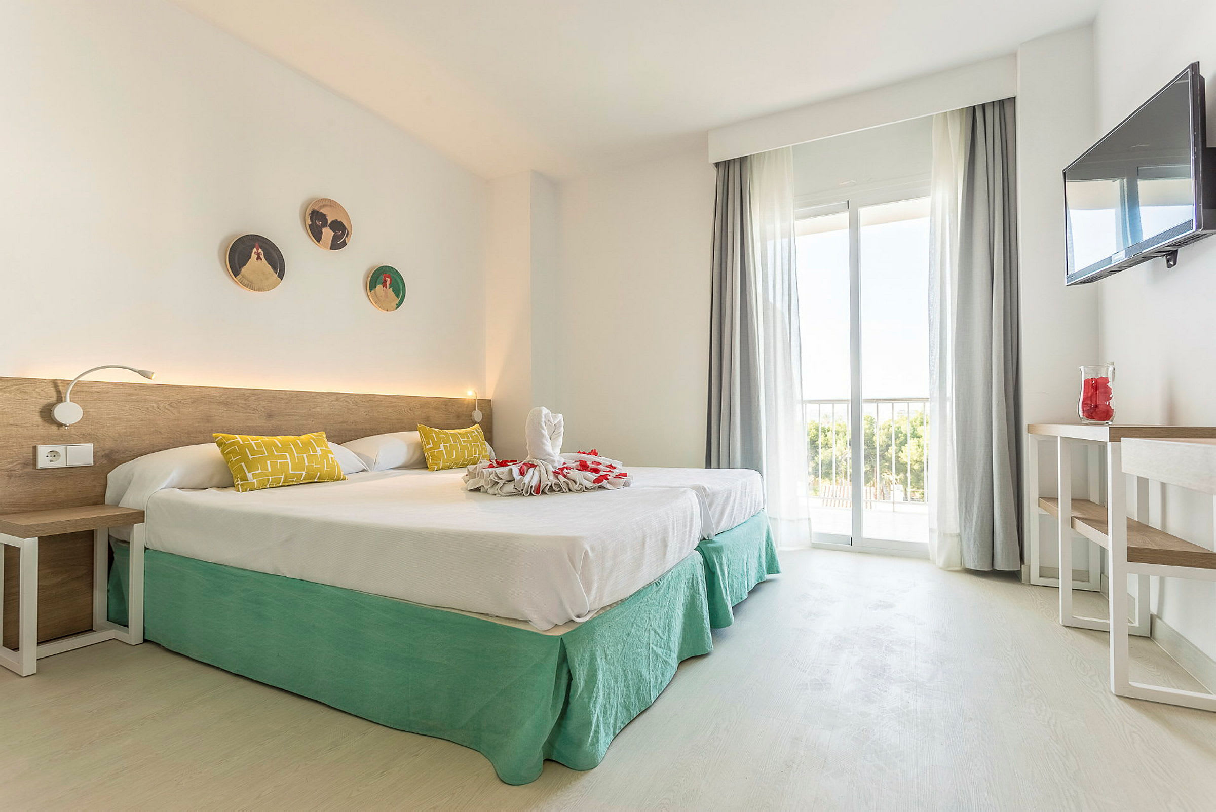 Et hotellrom med to enkeltsenger med hvitt sengetøy og grønne sengekapper. Sengen er dekorert med håndkleskunst og røde blomsterblader. Det er en hvit vegg med dekorasjoner, et skrivebord med en vase og en veggmontert TV. Rommet har store vinduer med lyse gardiner og utsikt over trær. (AI-generert tekst)