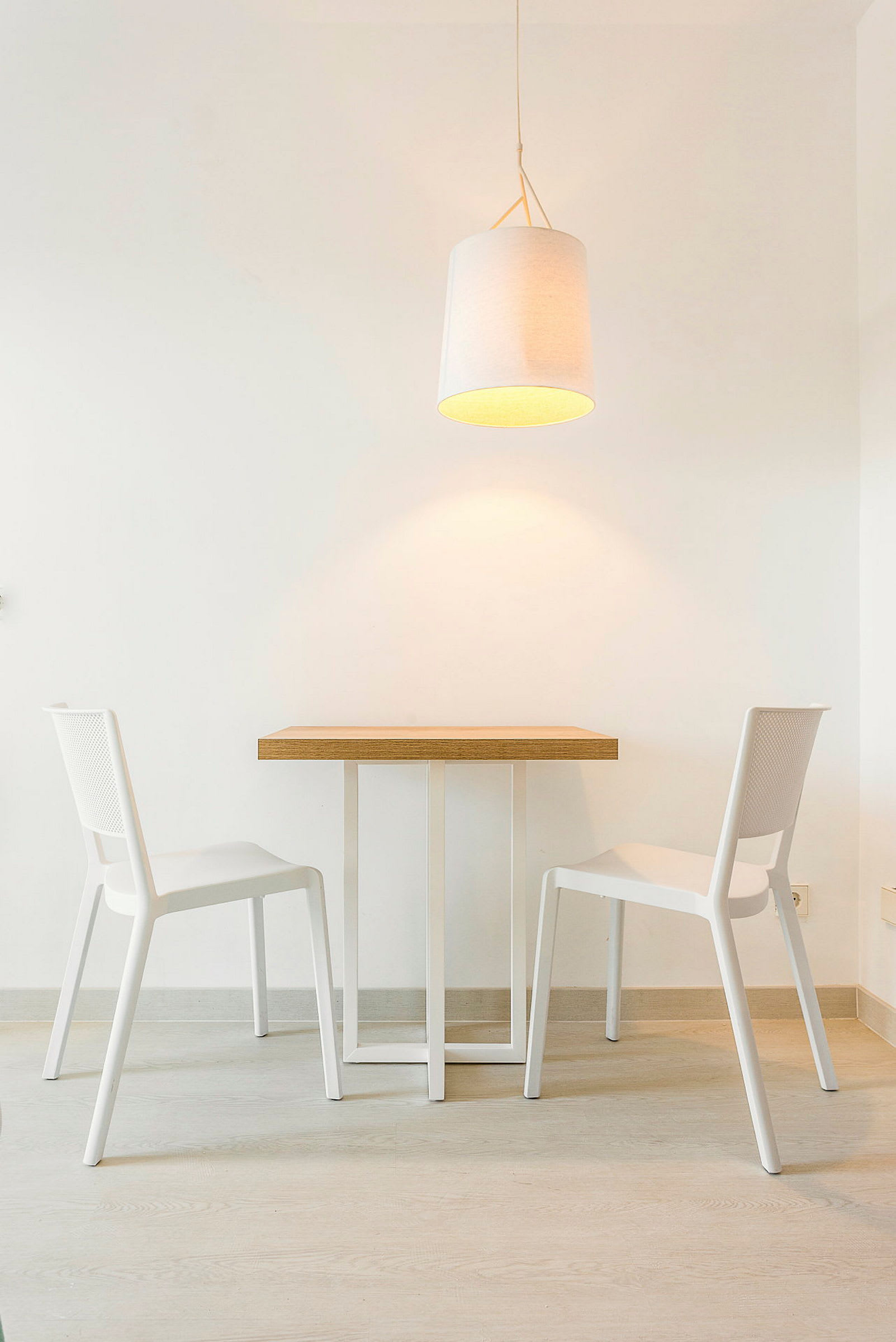 Et bord med to hvite stoler, og en hengende lampe over i et minimalistisk rom. (AI-generert tekst)
