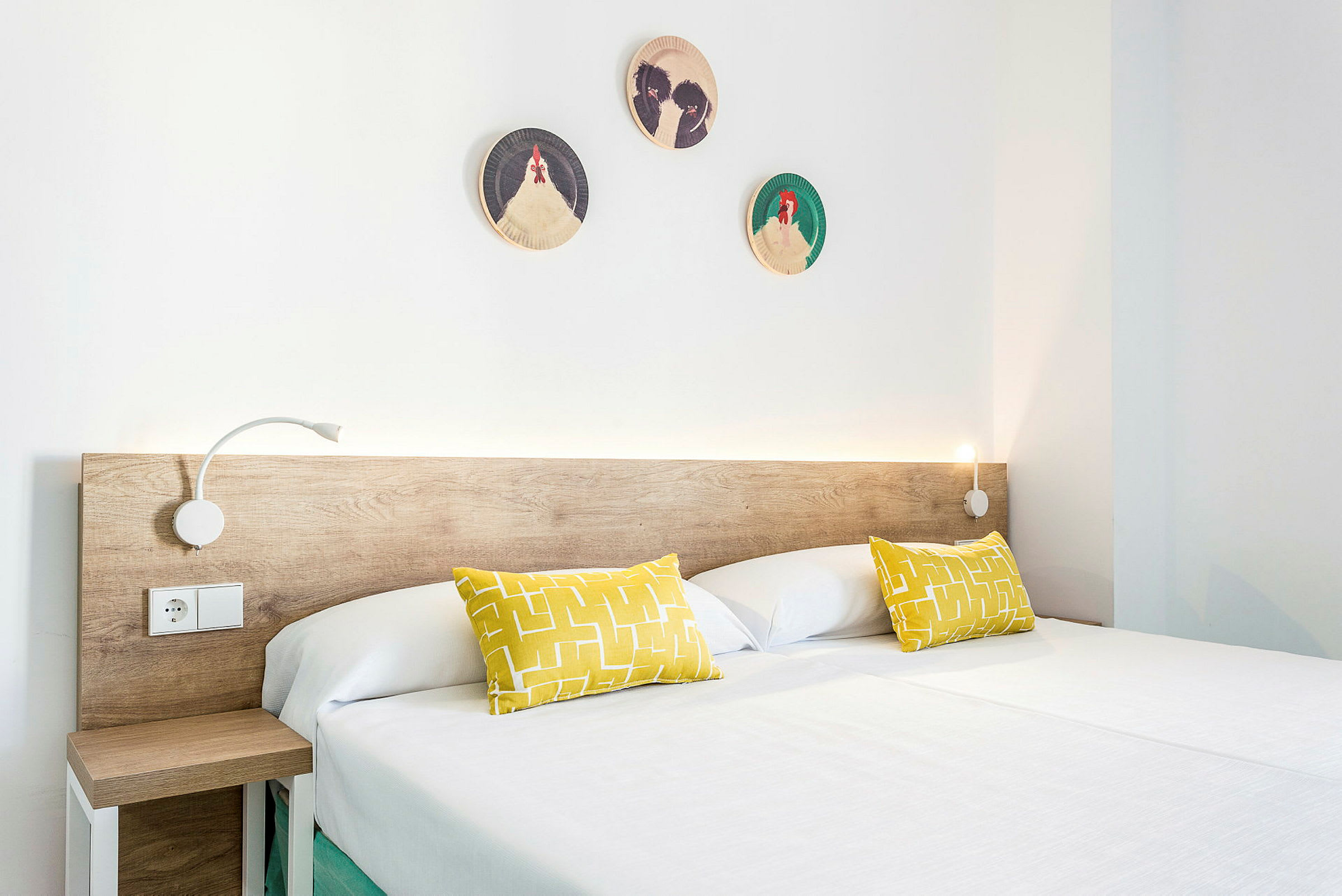 Et moderne hotellrom med en dobbeltseng. Sengen har en treramme, hvitt sengetøy og to gule puter. Veggdekorasjoner med fuglemønster er plassert over hodeenden. (AI-generert tekst)