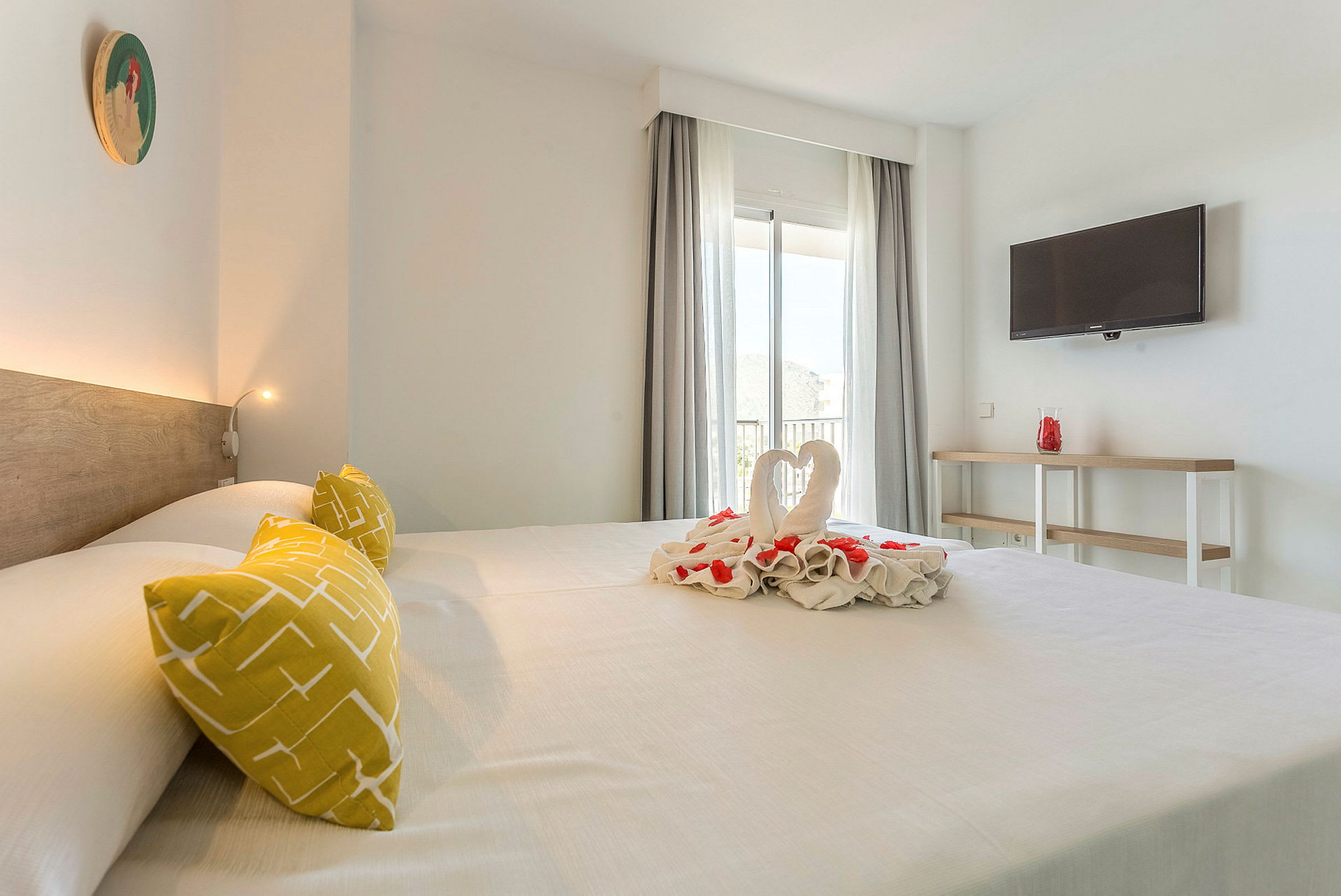Et hotellrom med en stor seng, dekorert med håndkledyr og blomster. Rommet har en TV på veggen og et vindu med gardiner. (AI-generert tekst)