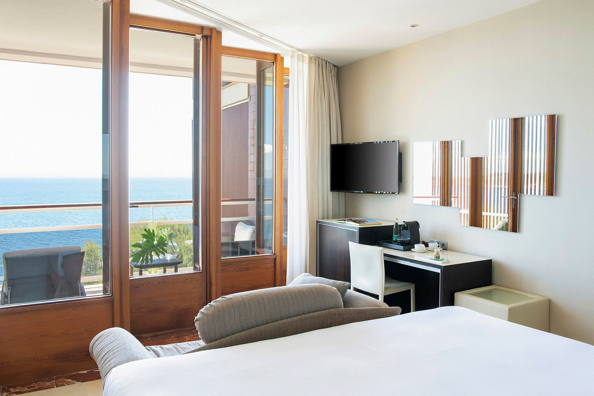 En moderne hotellromutsikt med en seng, skrivebord, TV og store glassdører som fører til en balkong med utsikt over havet. (AI-generert tekst)