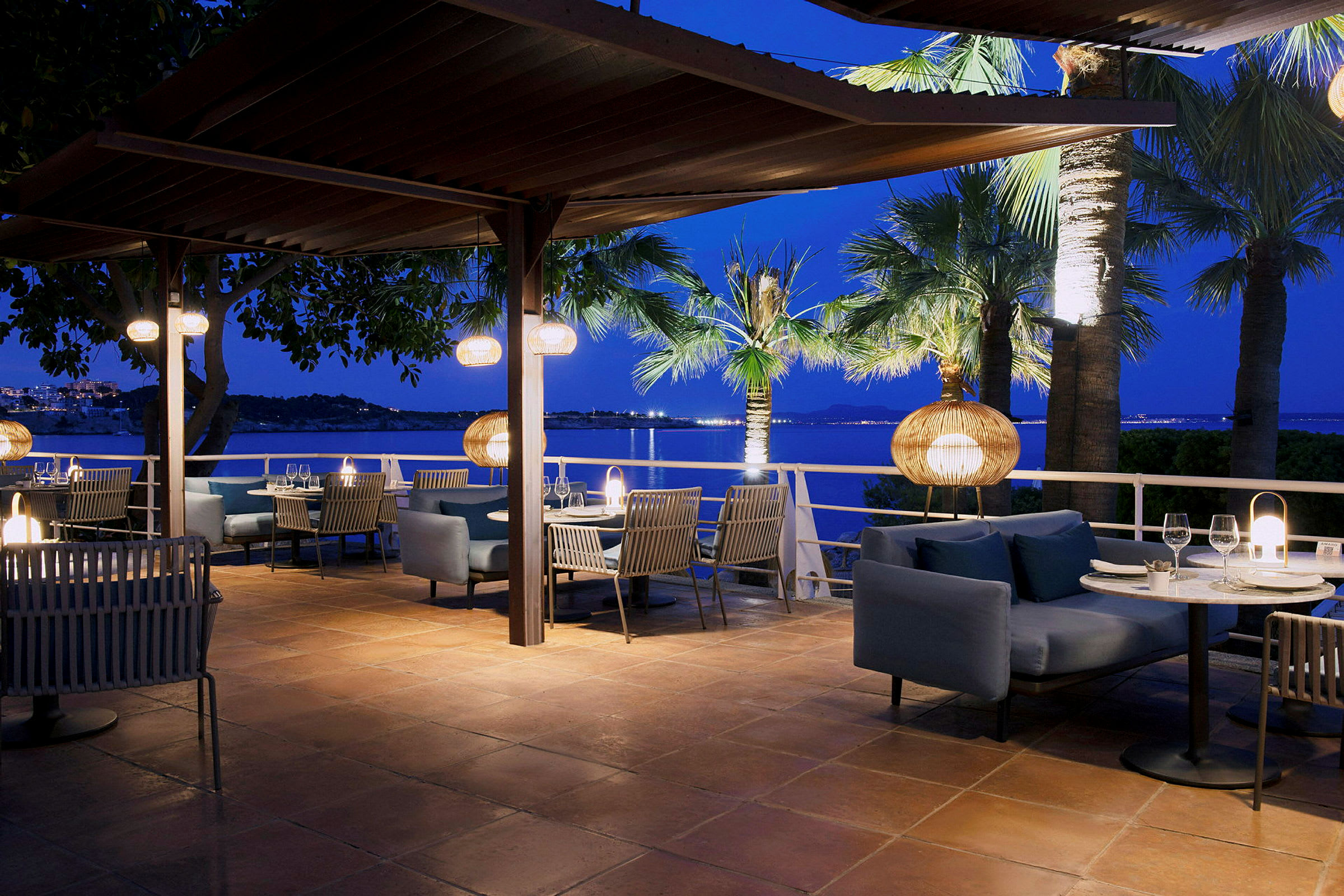 En restaurantterrasse om kvelden med bord og stoler, dekorert med hengende lamper og utsikt over blått vann og palmer. (AI-generert tekst)