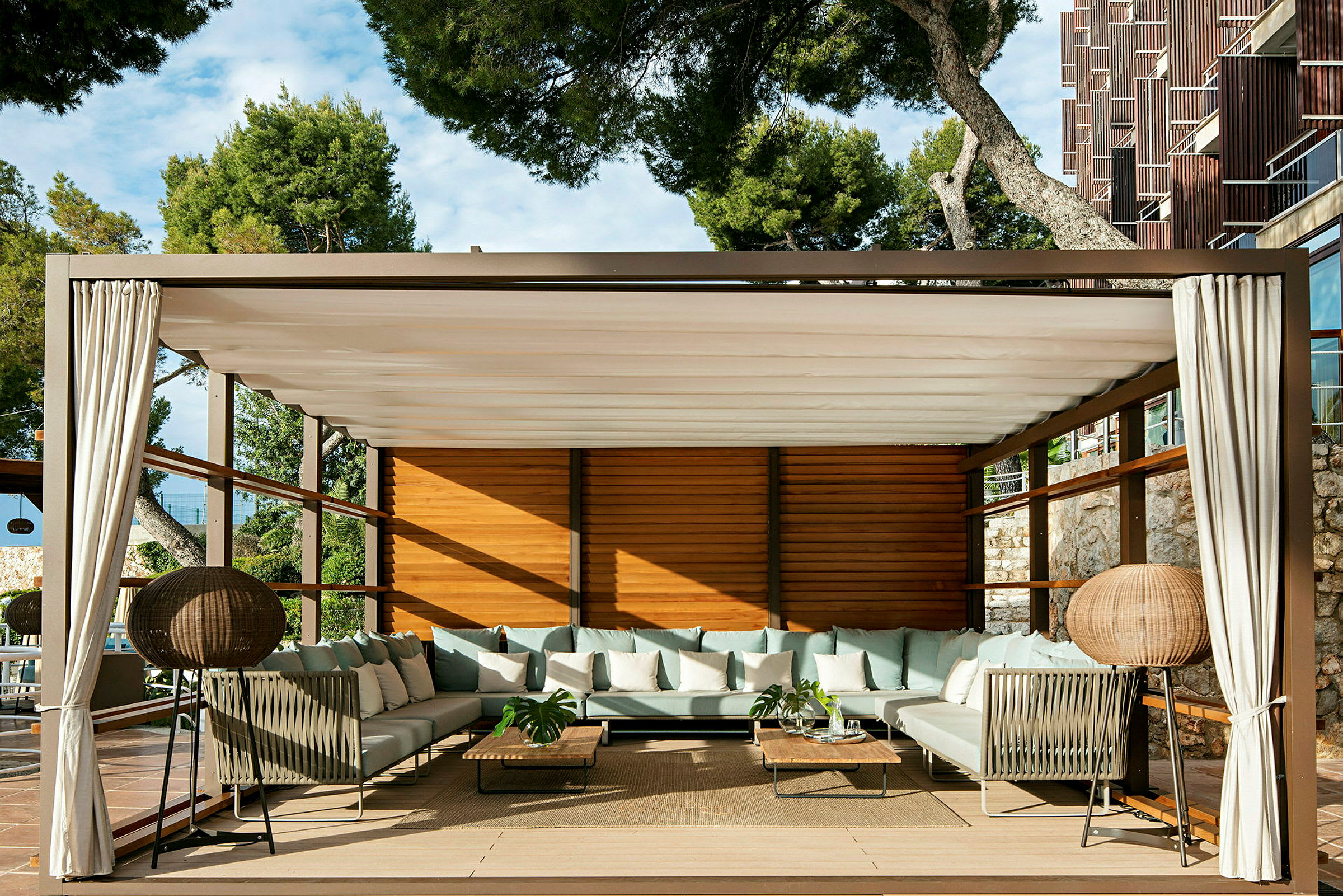 En lounge med sofaer, bord og gardiner under en pergola. (AI-generert tekst)