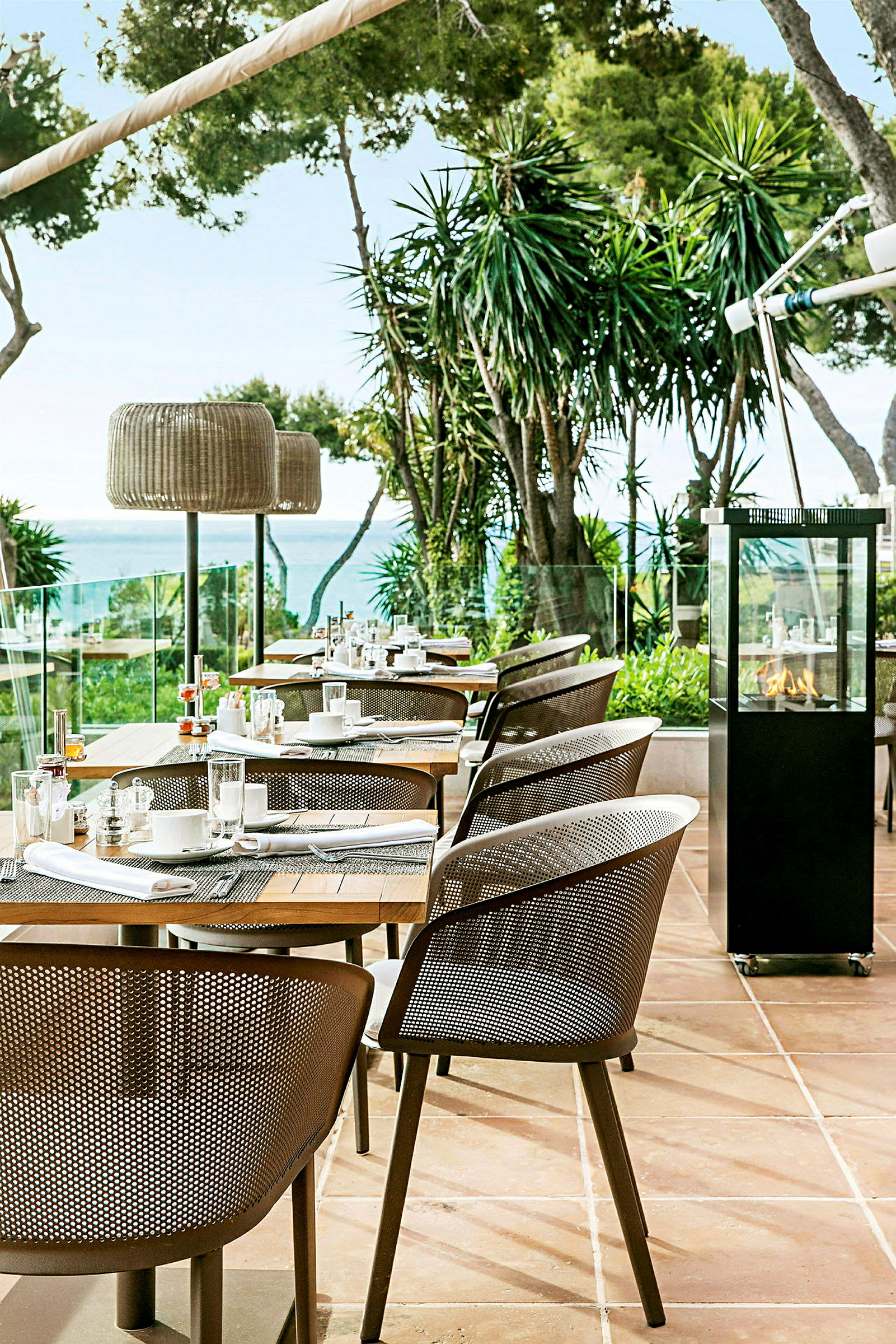 Utendørsrestaurant med moderne stoler og dekkede bord, omgitt av grøntarealer og utsikt mot havet. (AI-generert tekst)