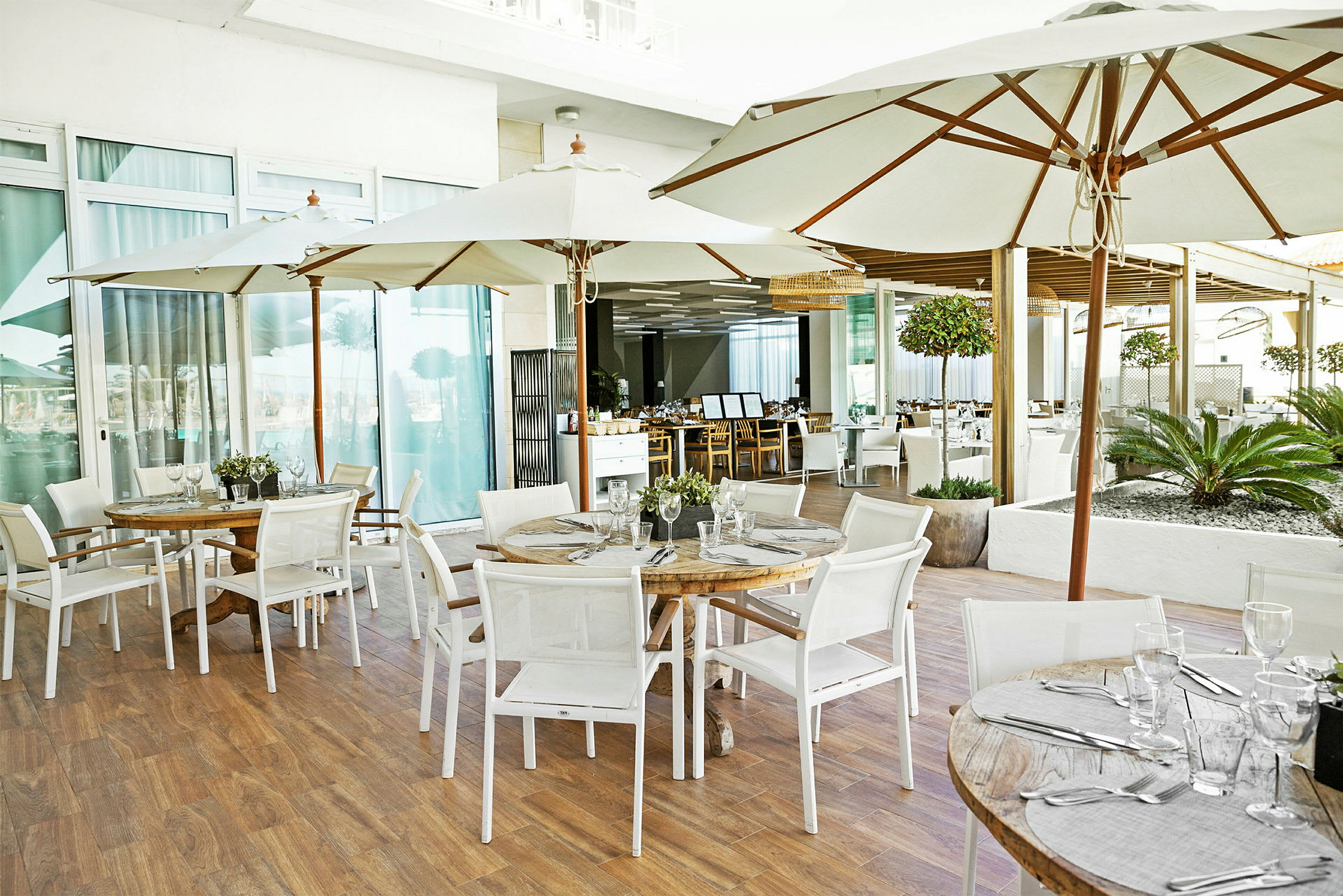 Bord med parasoller og stoler på en restaurantterrasse. Glass og bestikk ligger på bordene. (AI-generert tekst)