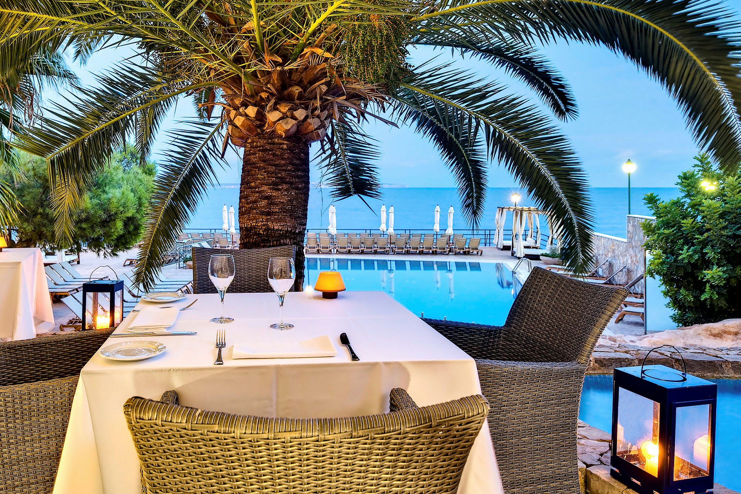 Utsikt over en restaurant ved et basseng nær havet med en palme i forgrunnen. (AI-generert tekst)