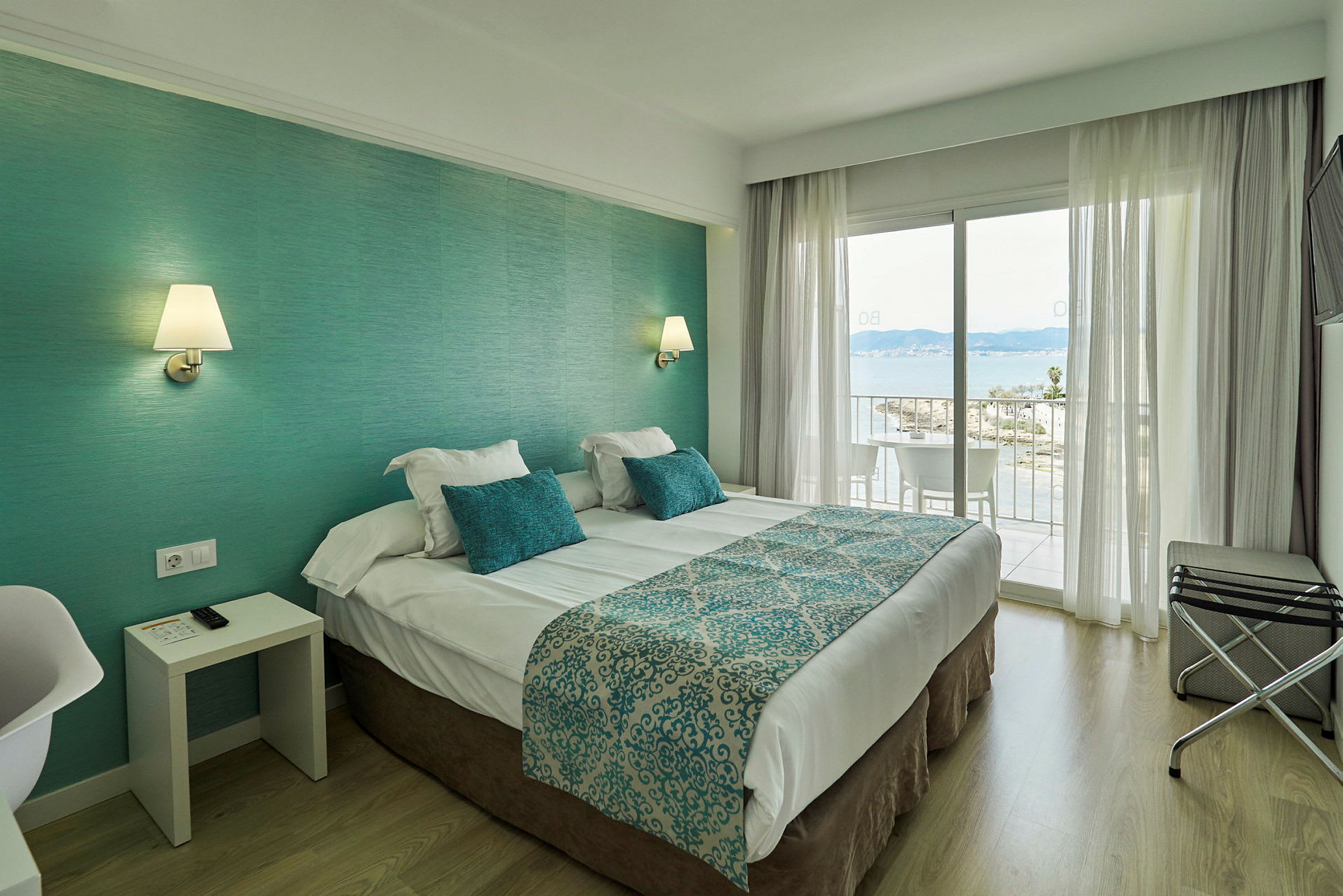 Et hotellrom med en dobbeltseng, turkist tema og utsikt over havet fra balkongen. (AI-generert tekst)