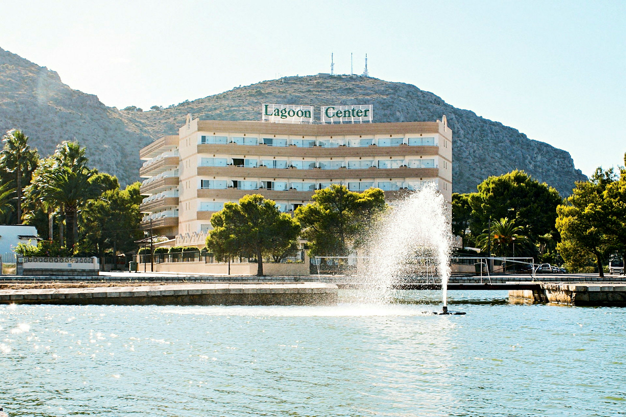 Hotell Lagoon Center ved strandpromenaden med en fontene og fjell i bakgrunnen. (AI-generert tekst)