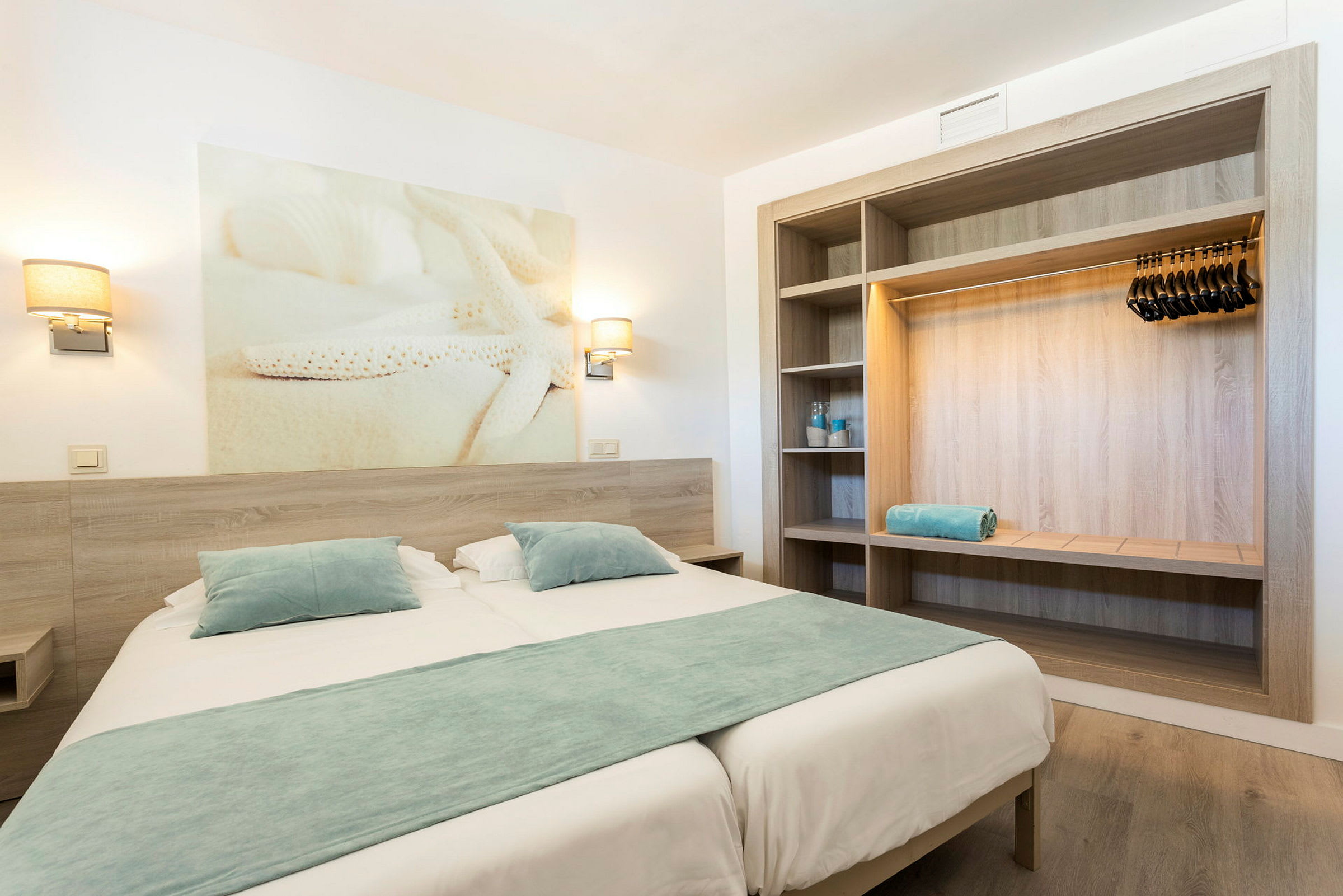Et hotellrom med en dobbeltseng, blå sengetøy, vegglamper og en åpen garderobe. (AI-generert tekst)