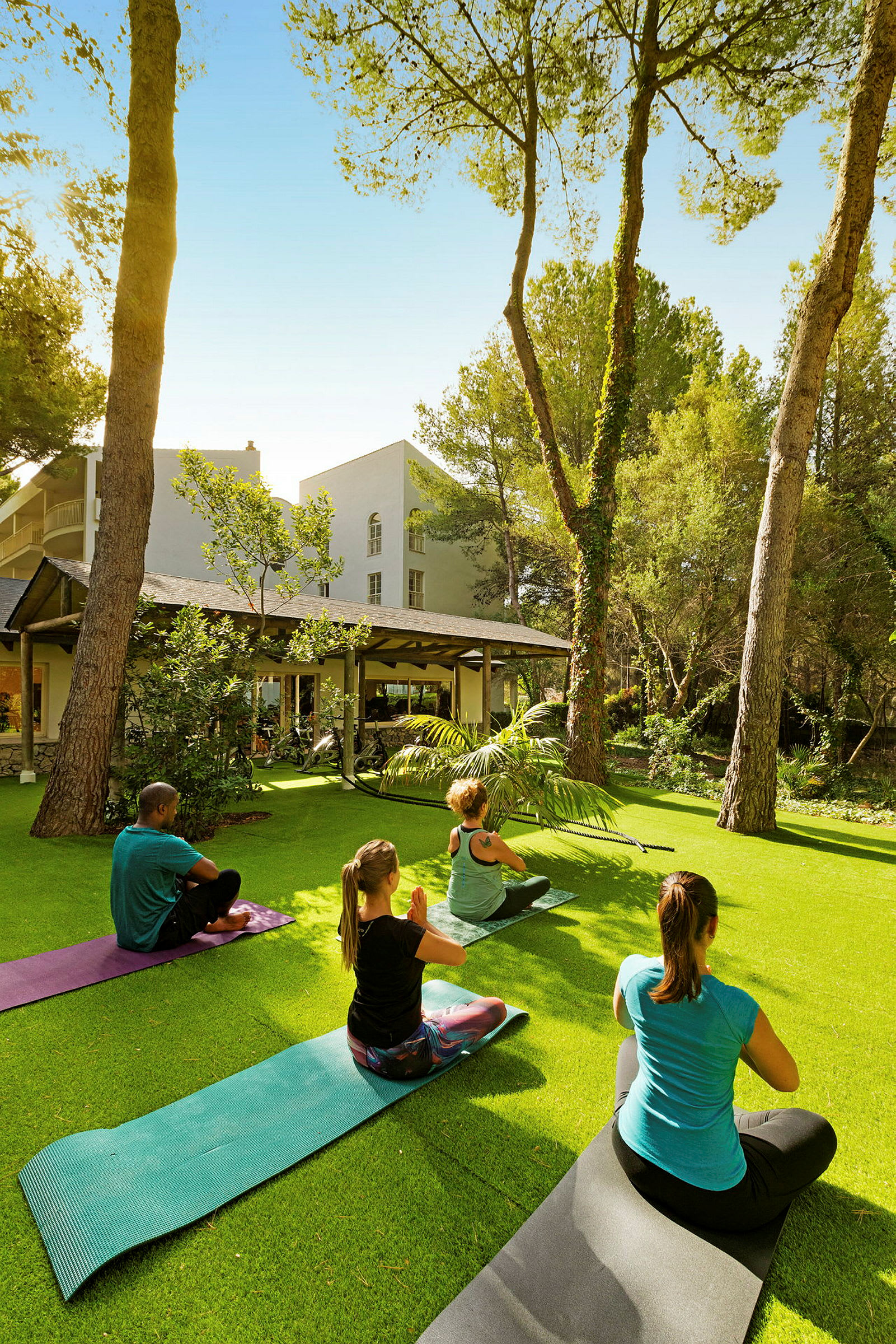 En gruppe personer praktiserer yoga utendørs på plenen foran bygninger omgitt av trær. (AI-generert tekst)