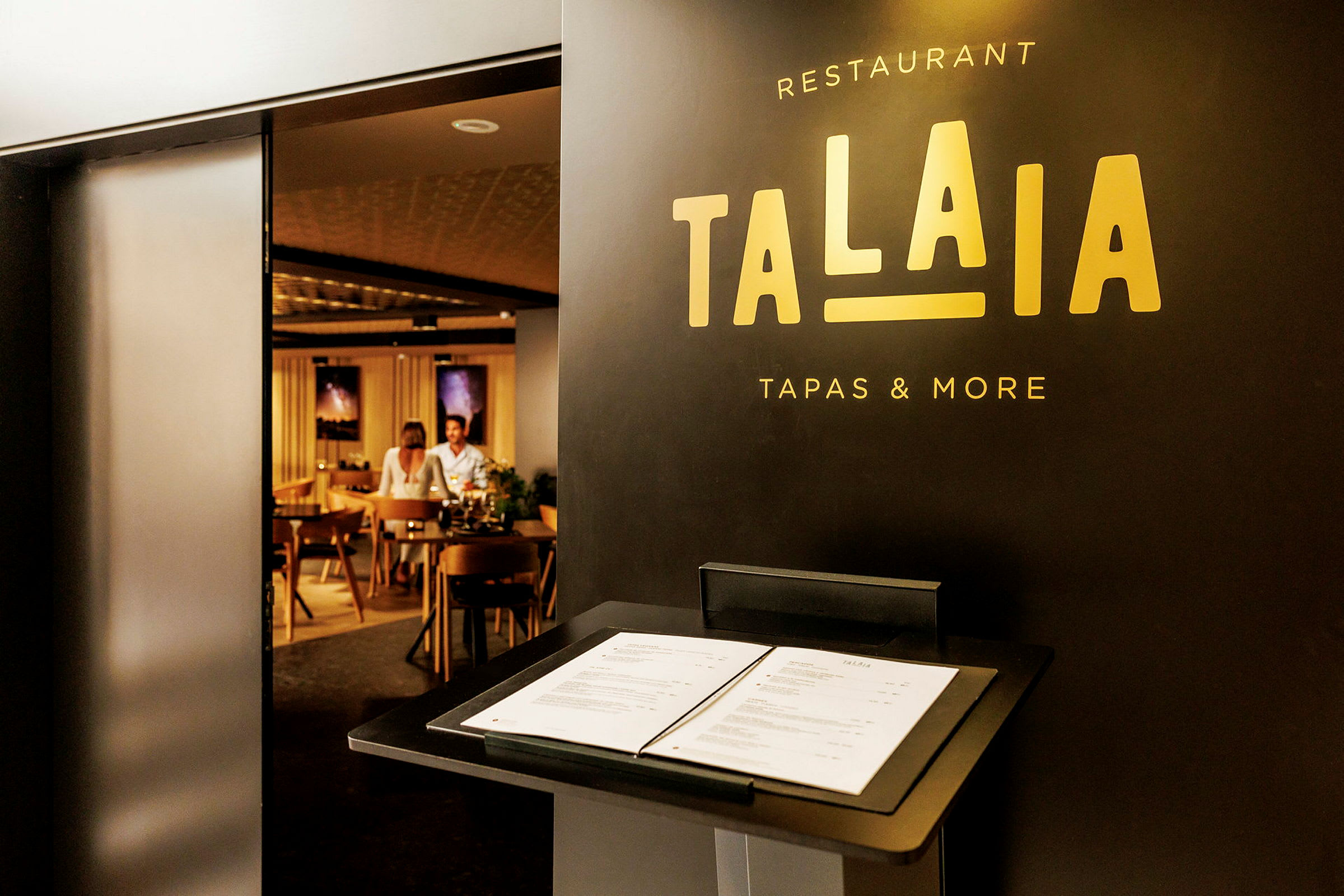 Restaurant Talaia med en meny på en stand ved inngangen, som viser et glimt inn i en stilfull interiør med sitteplasser. (AI-generert tekst)