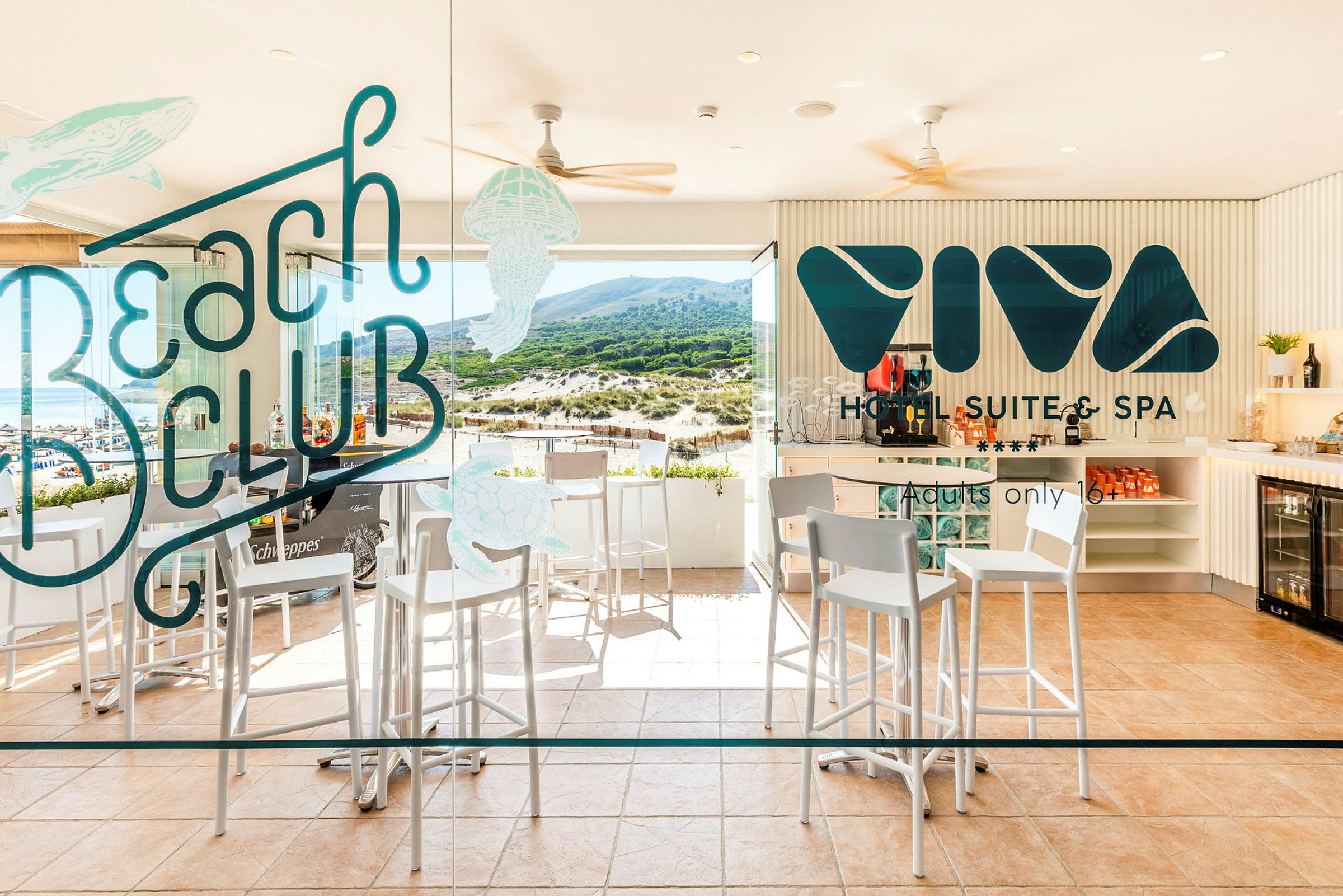 Bilde av en hotellbar med høye stoler, bord og glassvegg med teksten 'Beach Club' og 'Viva Hotel Suite & Spa' samt utsikt mot en strand og grønne høyder. (AI-generert tekst)