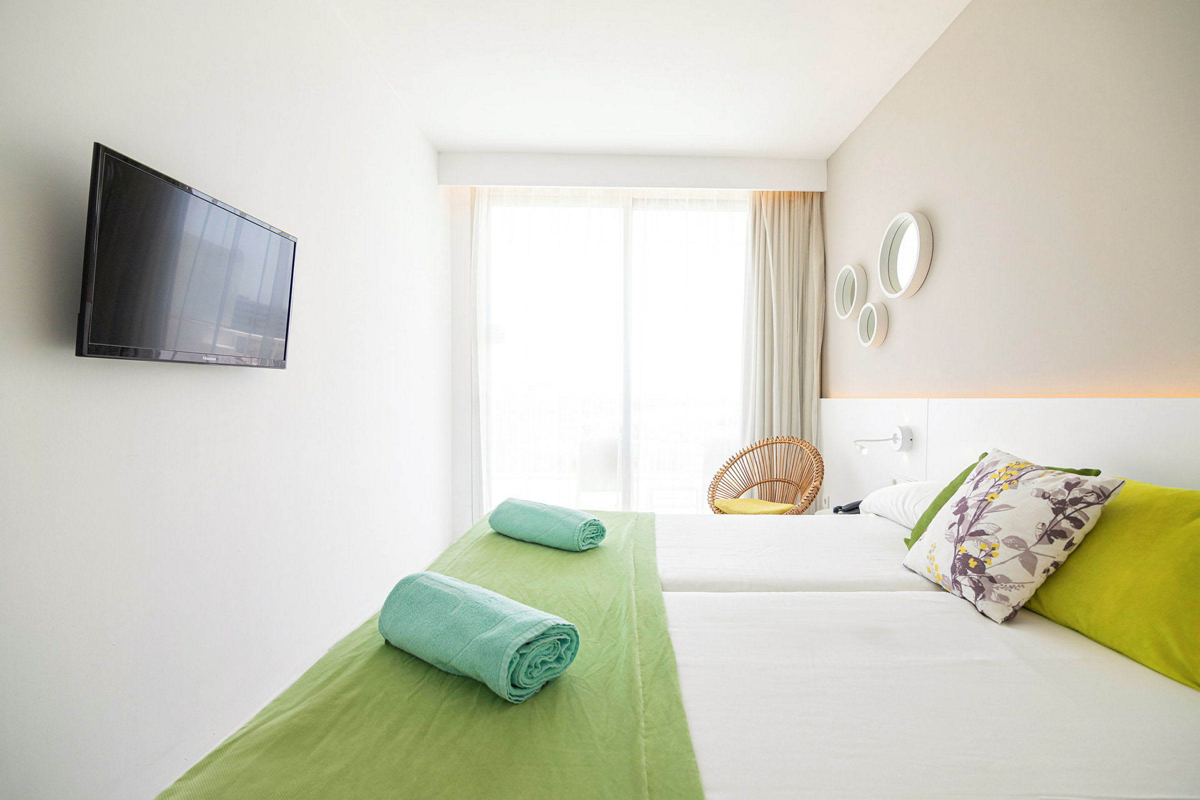 Et lyst hotellrom med en dobbeltseng, fargerike puter, sammenrullede håndklær og en flatskjerm-TV på veggen. (AI-generert tekst)