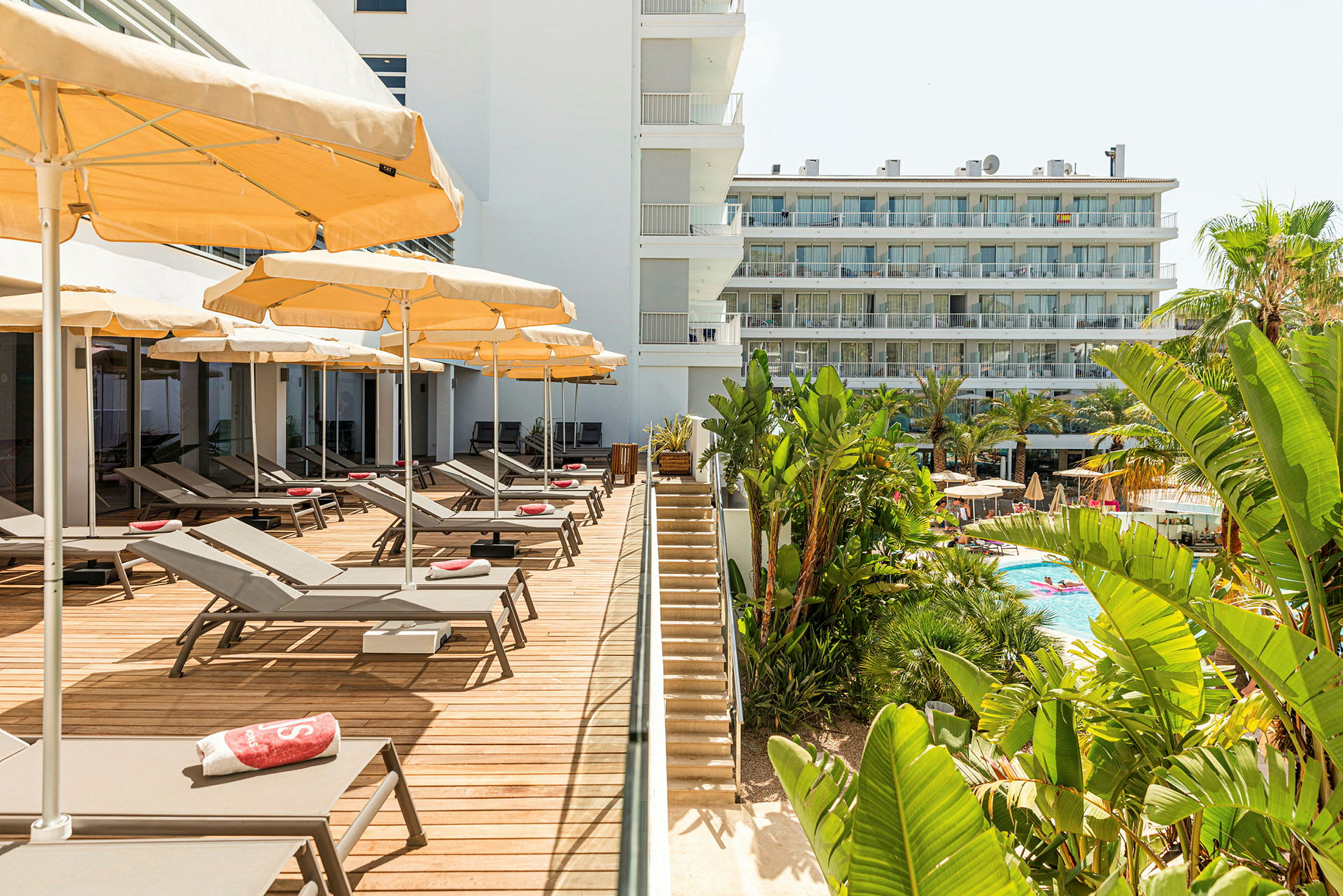 Et hotellområde med liggestoler og parasoller på en terrasse med tredekke ved siden av en trapp som fører til bassengområdet, omgitt av frodig vegetasjon. (AI-generert tekst)