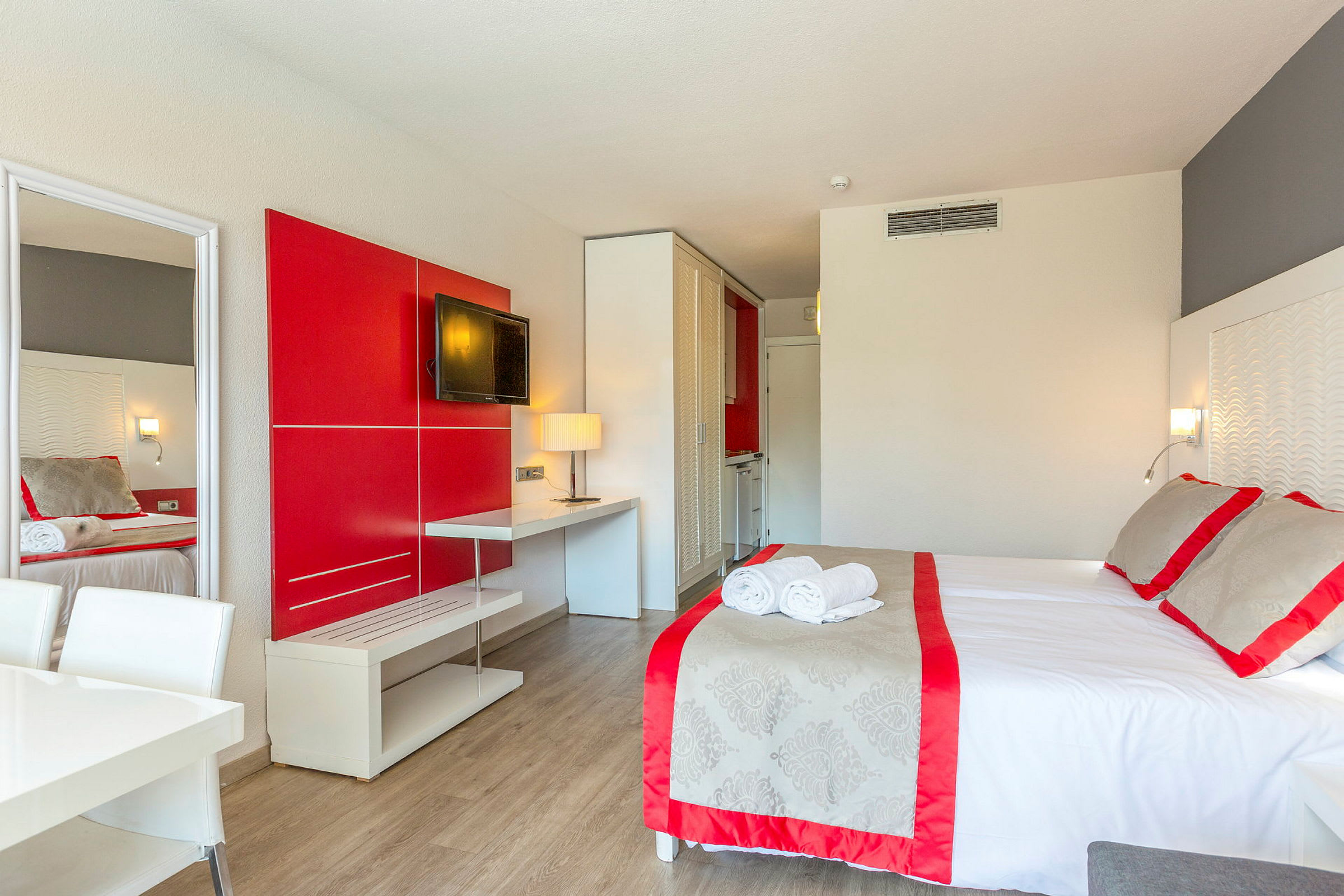 Et hotellrom med en seng, røde og grå detaljer, en TV på veggen og et bord med en lampe. (AI-generert tekst)
