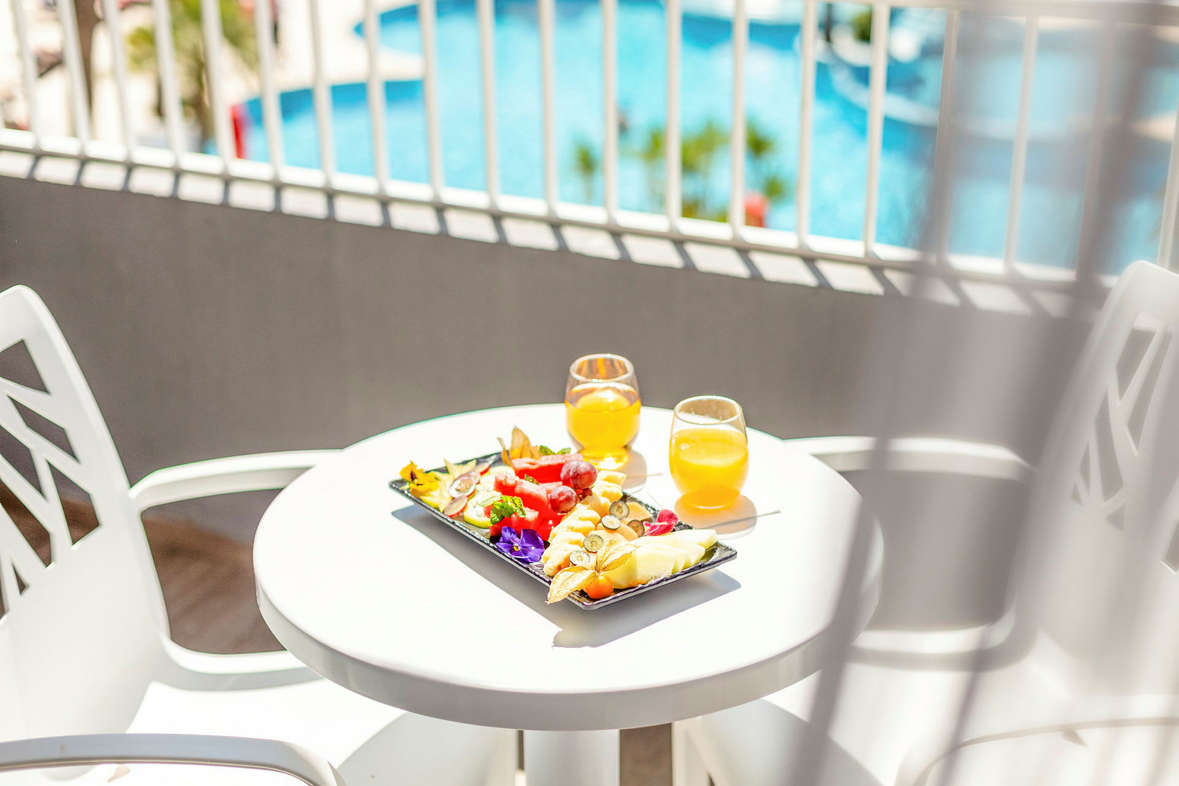 Et hvitt bord med frisk frukt og to glass appelsinjuice, med et basseng i bakgrunnen. (AI-generert tekst)