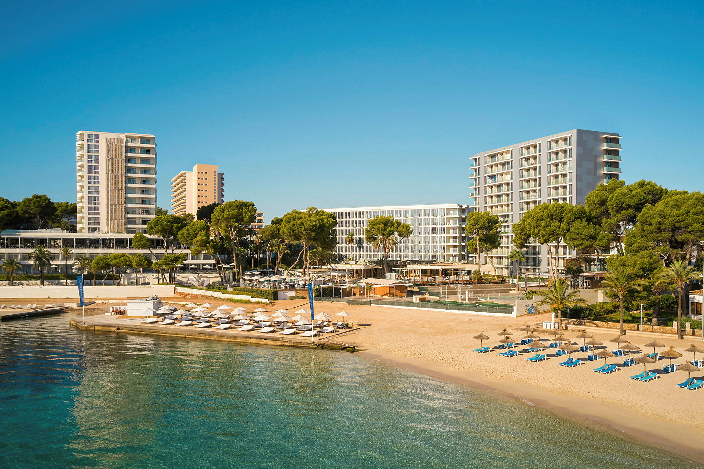 En strand med solsenger og parasoller ved siden av hotellbygninger og grøntområder. (AI-generert tekst)