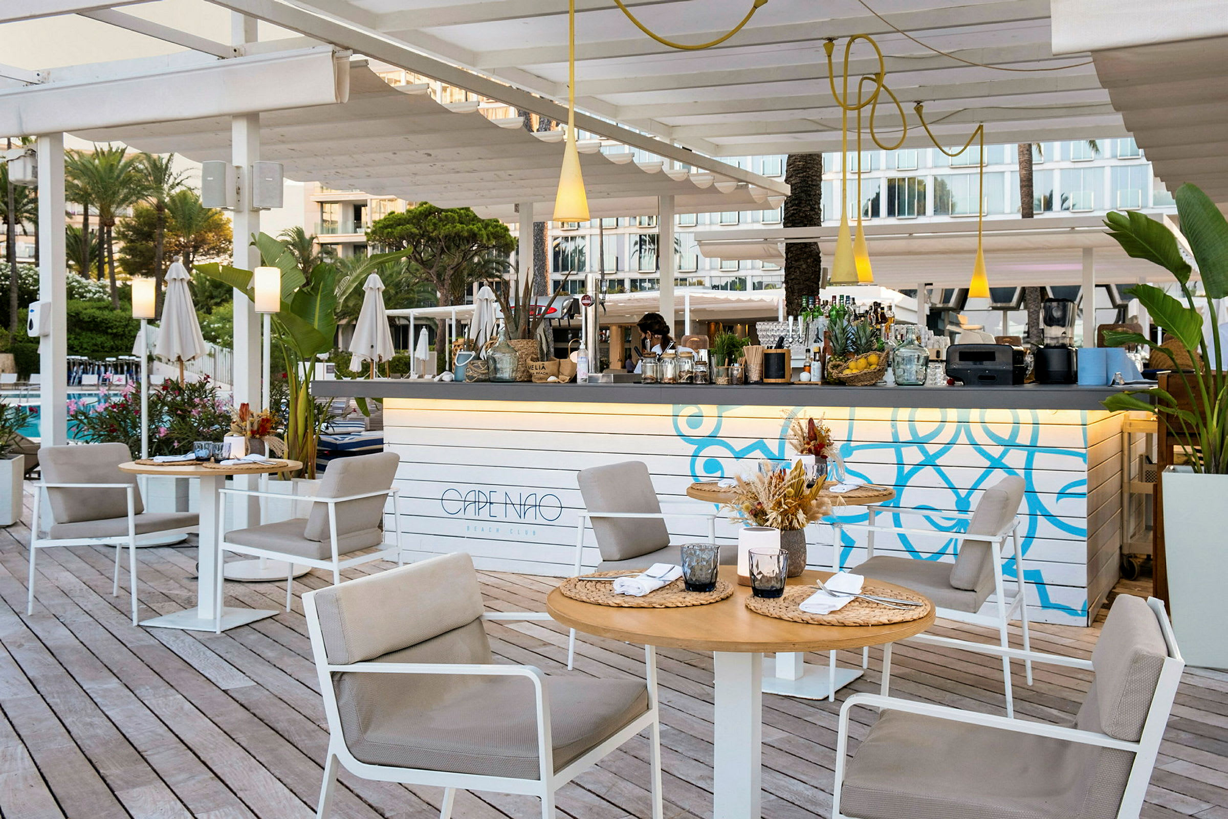 Utendørsmiljø ved en strandklubb med sittegrupper, et dekorativt barområde og grønne planter. (AI-generert tekst)