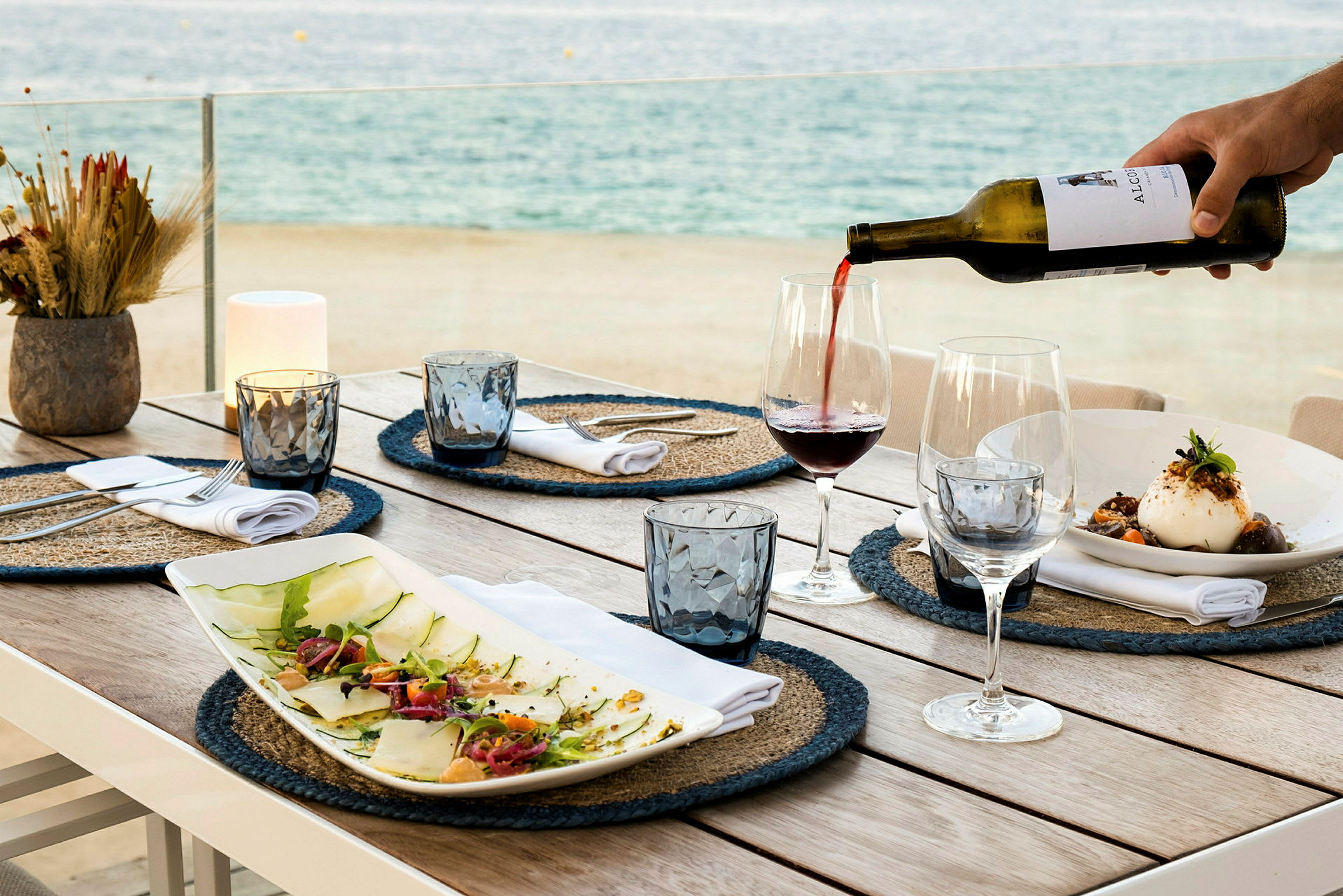 Et dekket bord med tallerkener, glass og flasker. Vin helles opp i et glass med havet i bakgrunnen. (AI-generert tekst)