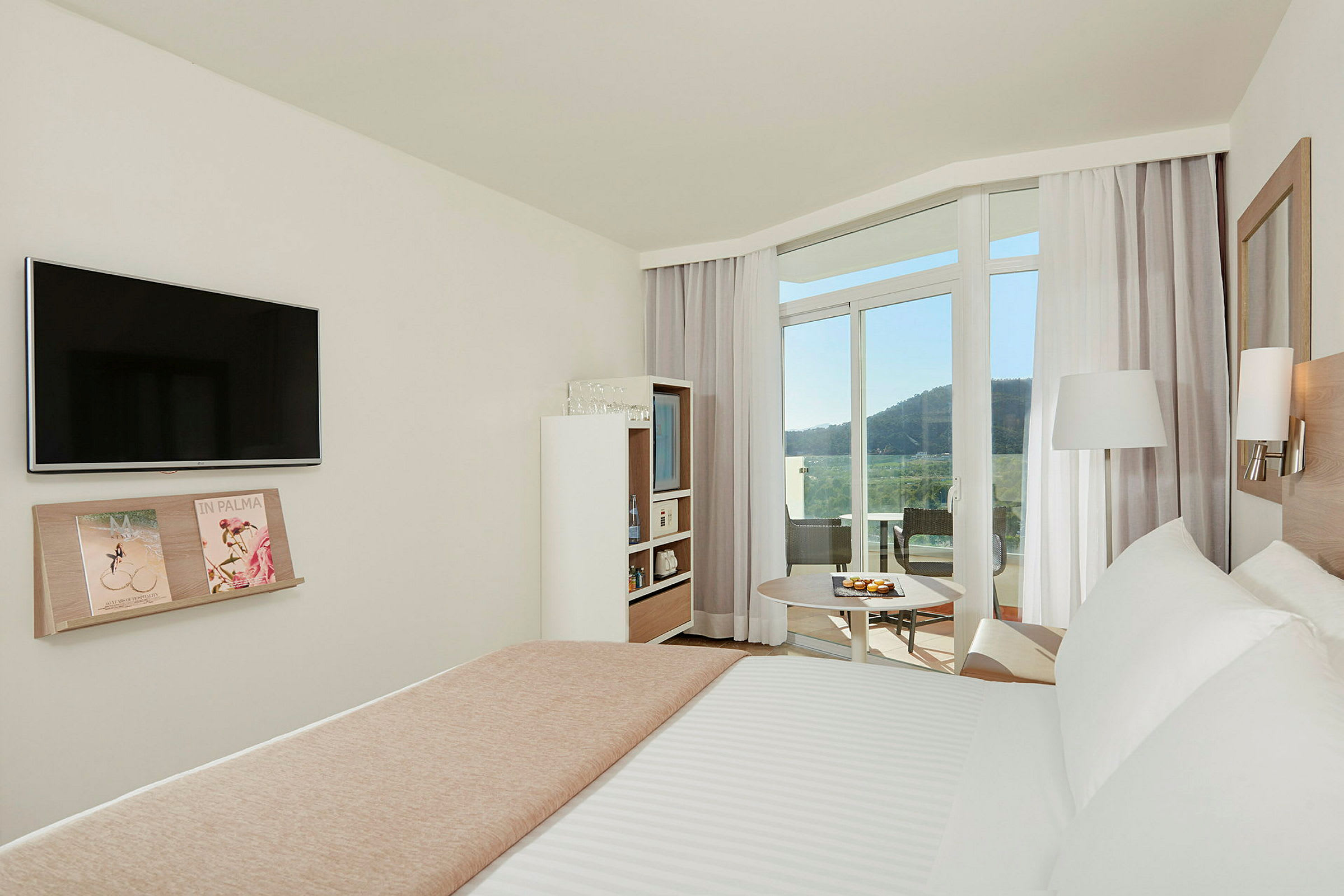 Et lyst og moderne hotellrom med en dobbeltseng, TV på veggen og en hylle med magasiner. Rommet har store vinduer og en balkong med utsikt over et frodig fjellandskap. (AI-generert tekst)