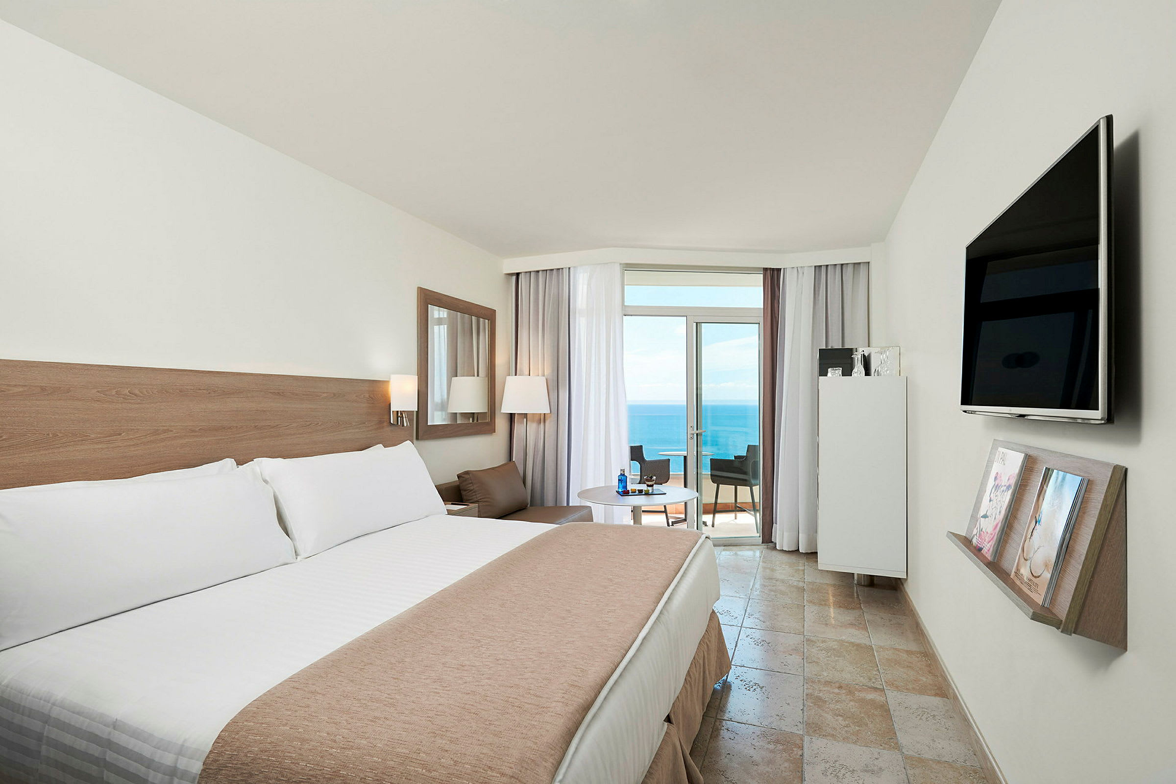 Hotellrom med en stor seng, et bord med stoler og en balkong med utsikt over havet. (AI-generert tekst)